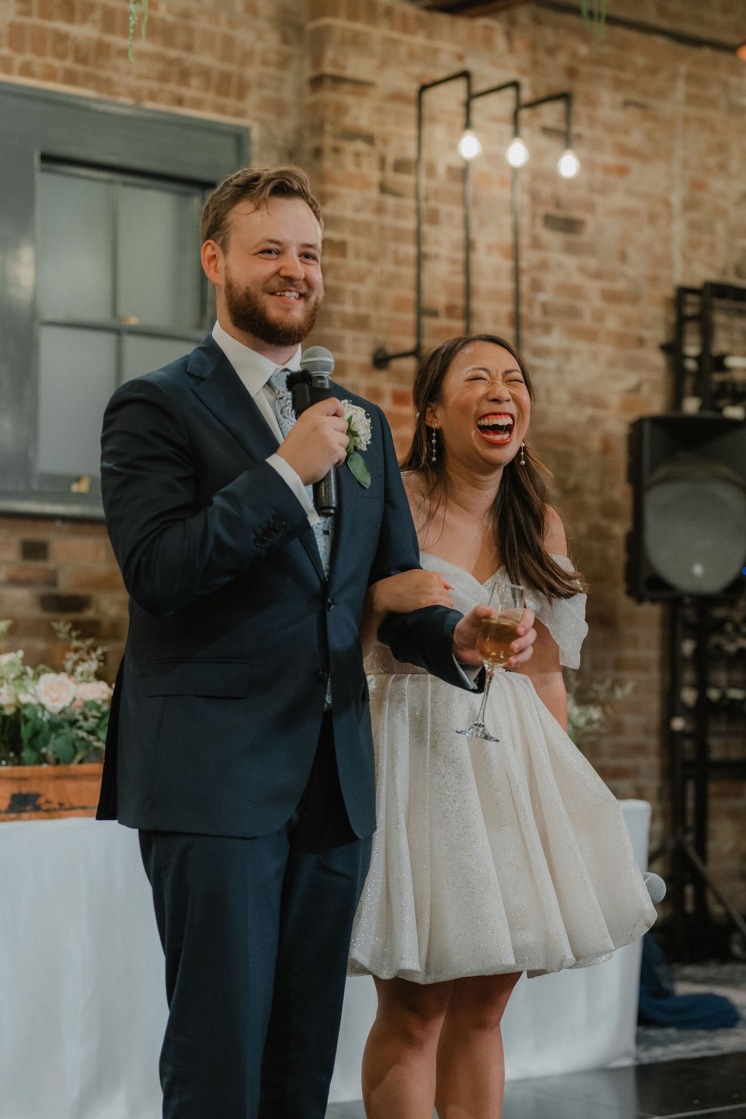 Couple laughs together while giving wedding speeches in front of exposed brick walls.