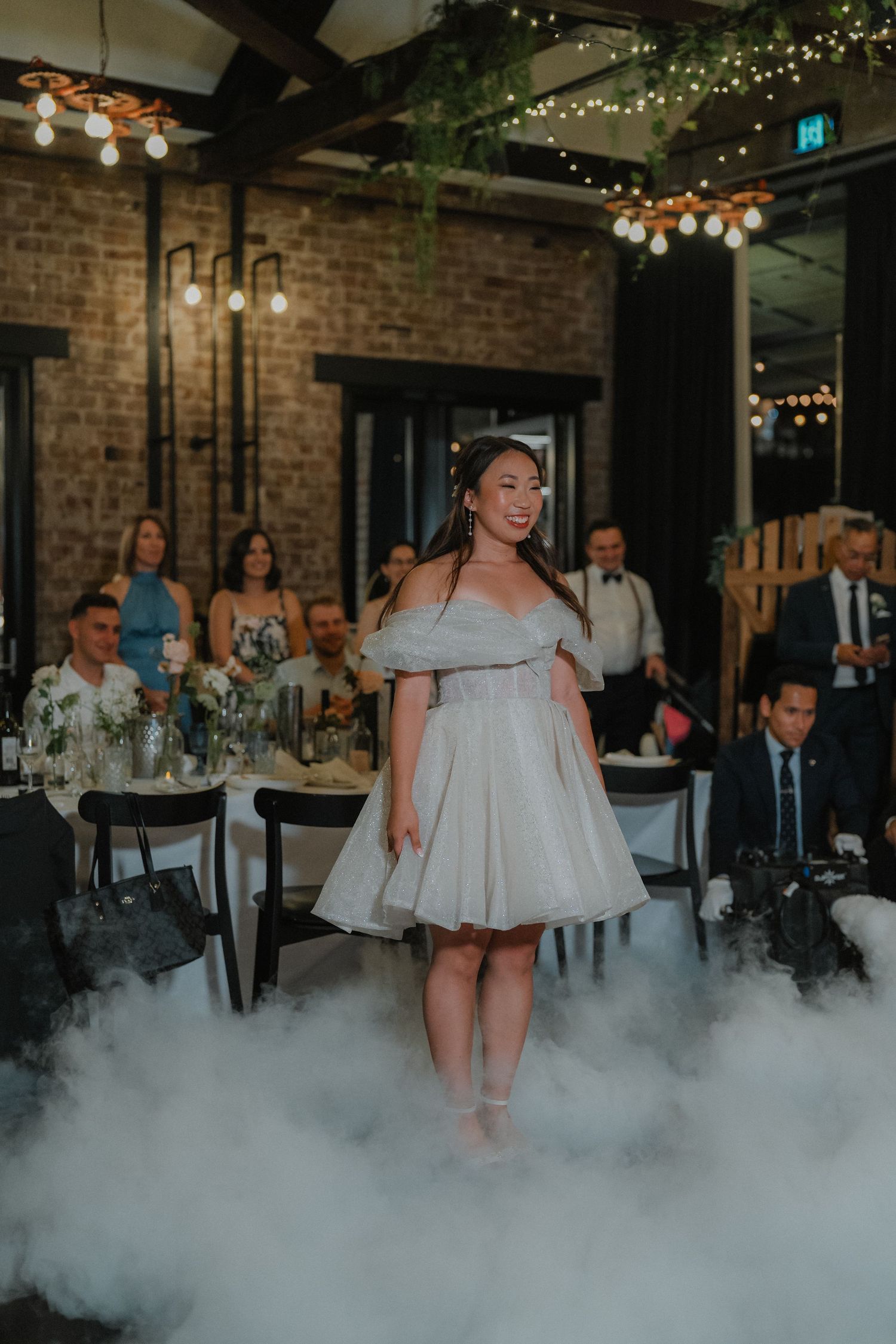 Bride stands in fog effect during reception in an industrial-style venue with string lights.