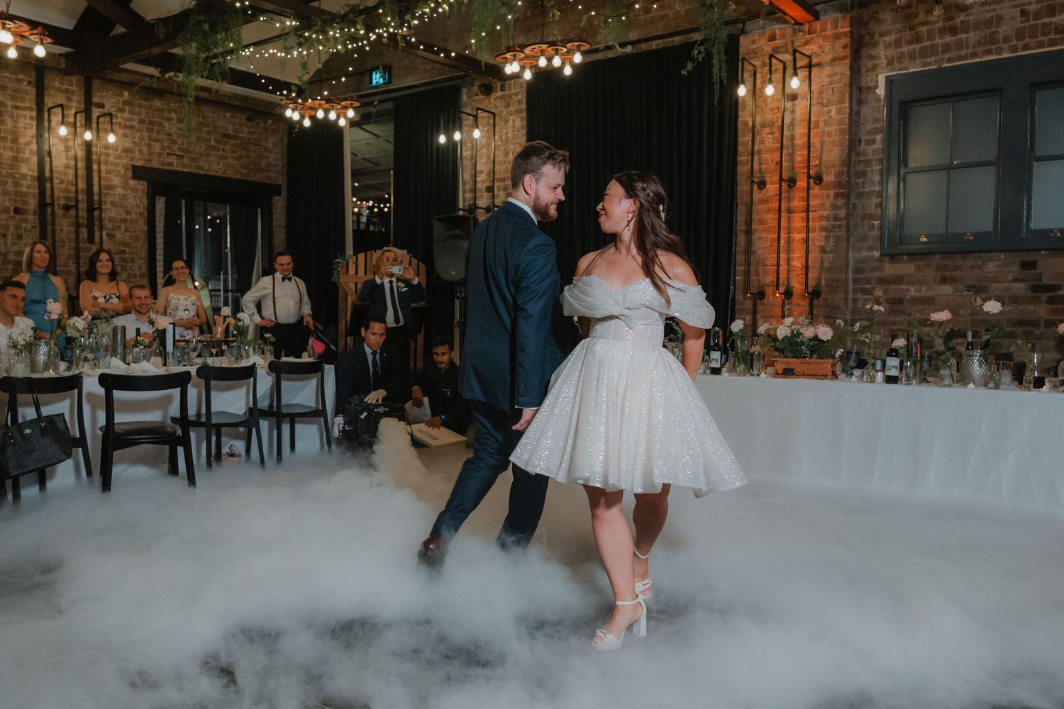 Wedding reception venue features exposed brick walls, string lights, and white decor in an industrial chic setting.