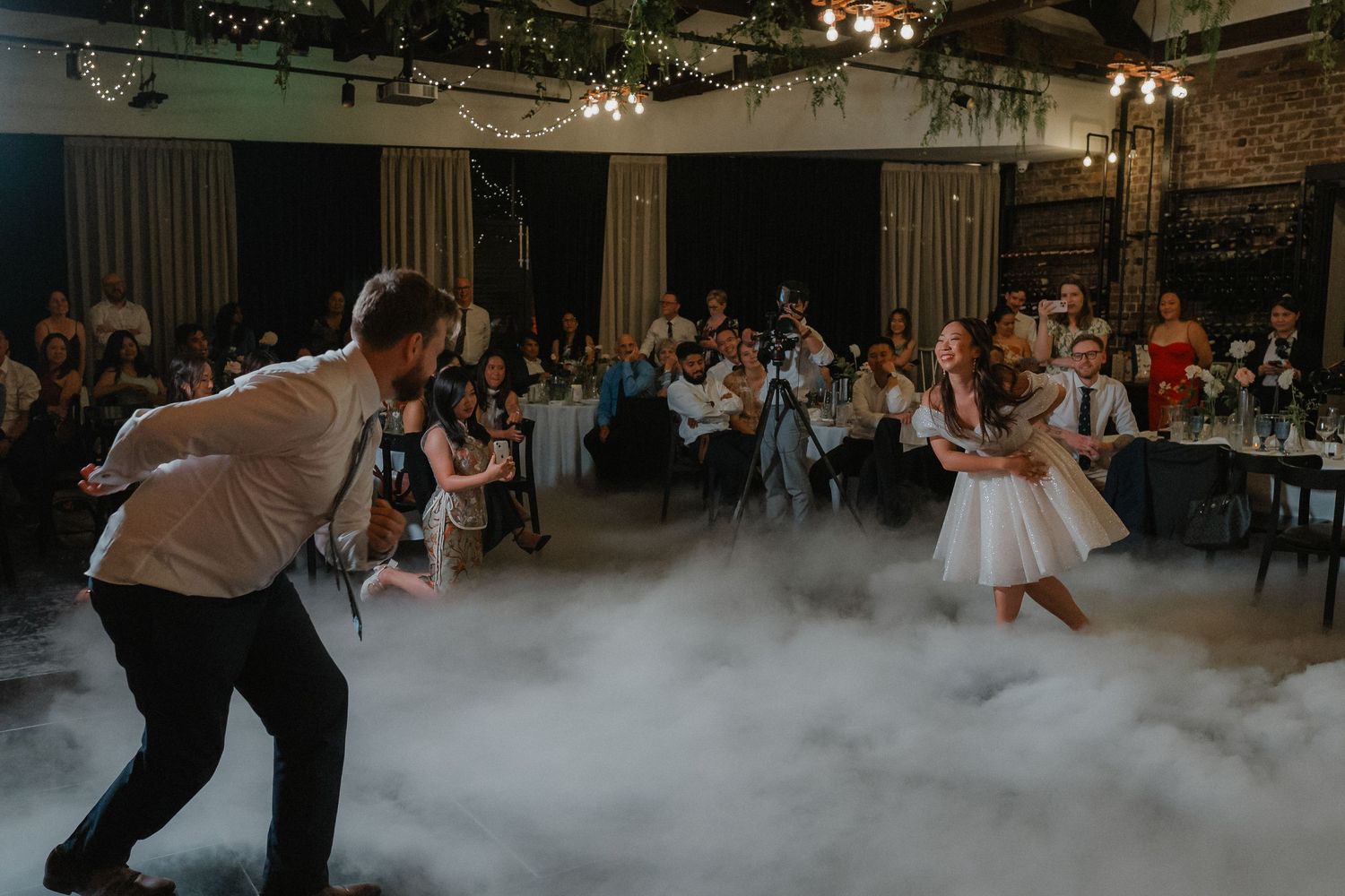 Wedding guests dance on a fog-filled dance floor in an industrial-style venue with exposed brick walls.
