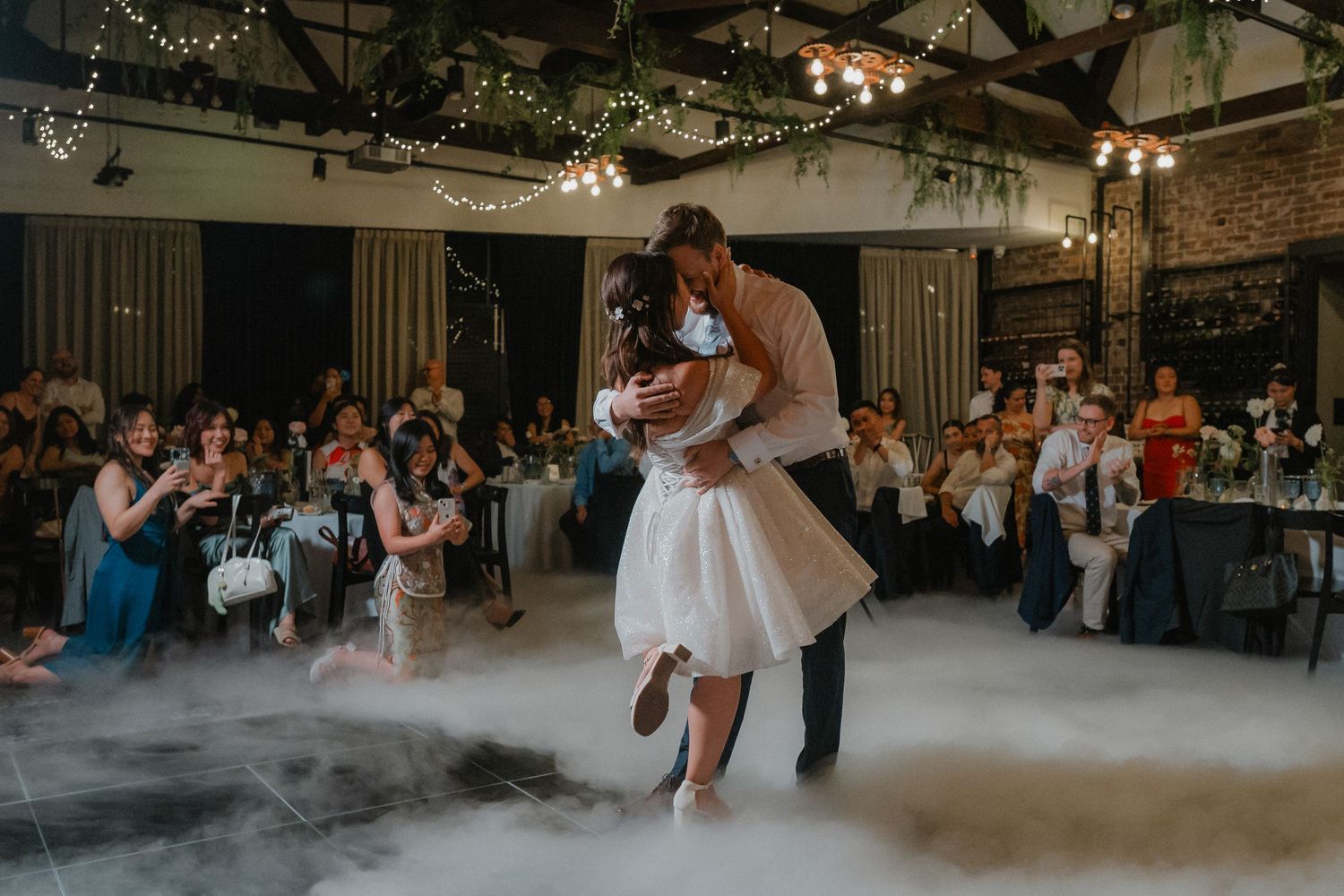 Couple dances in fog during their first dance while guests watch from reception tables.