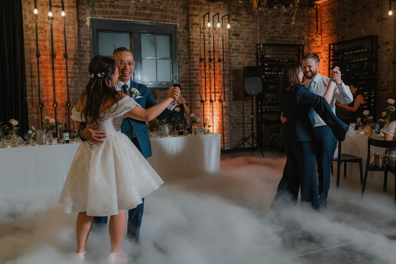 Wedding dance floor filled with low fog effects in industrial brick venue with ambient lighting.