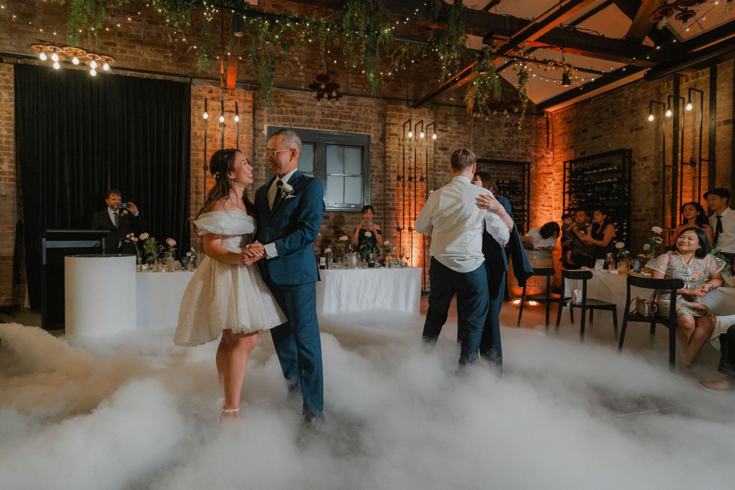 Guests dance beneath twinkling lights and greenery in a rustic brick venue with atmospheric fog.