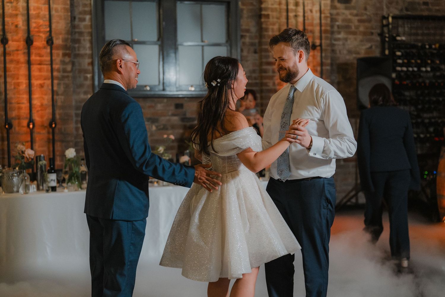 Couples dancing in a dimly lit industrial venue with warm orange lighting and exposed brick walls.