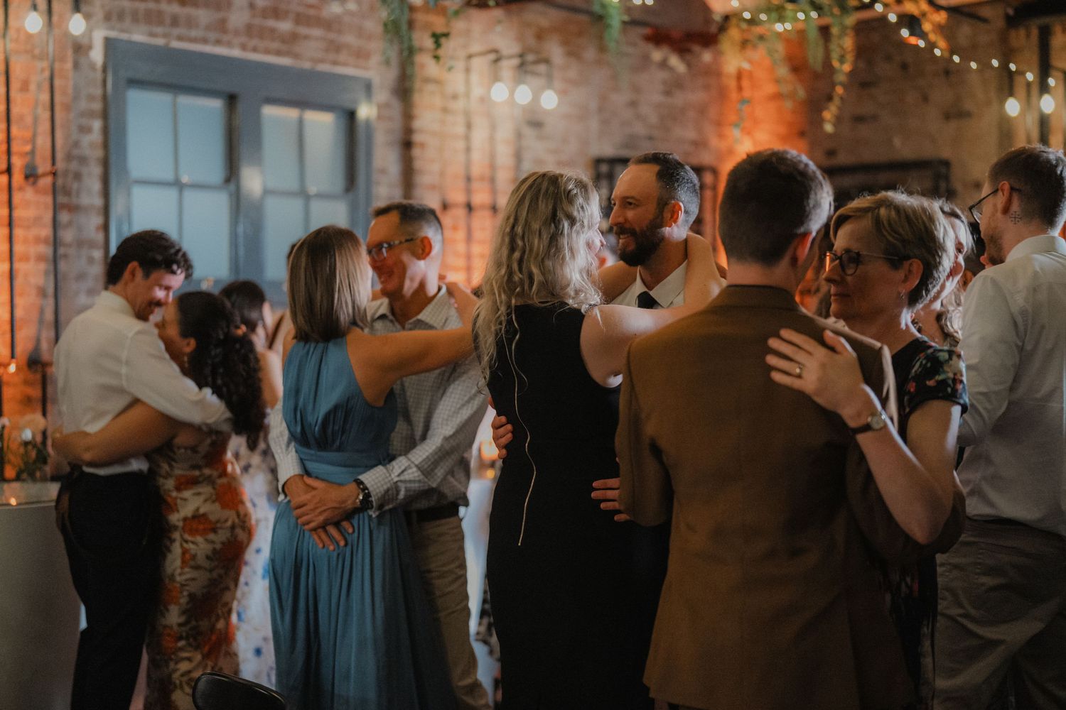 Couples slow dancing in dimly lit industrial venue with exposed brick walls and string lights.