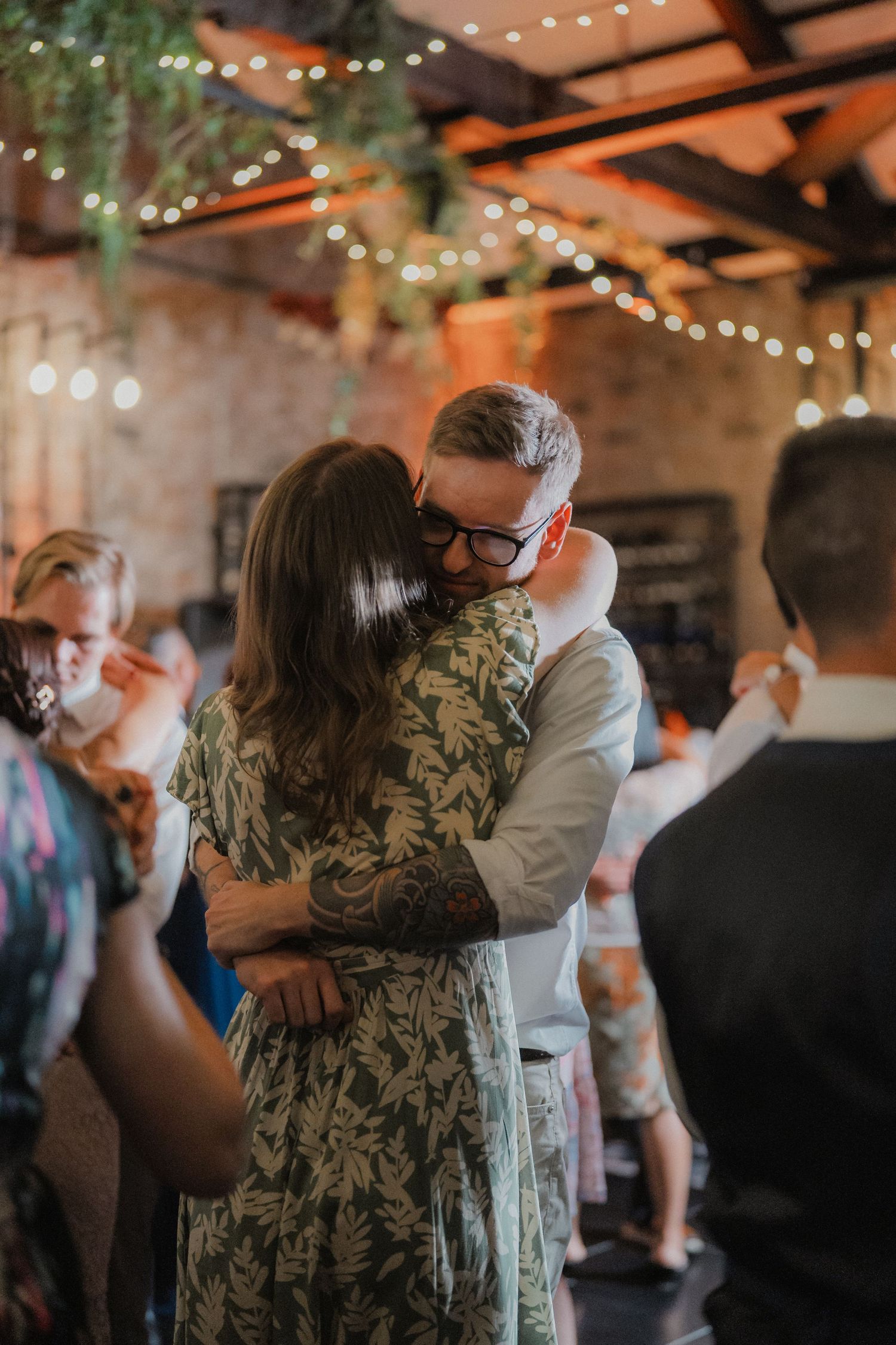 Wedding guests dancing and hugging on the dance floor under romantic string lights in a rustic barn venue.