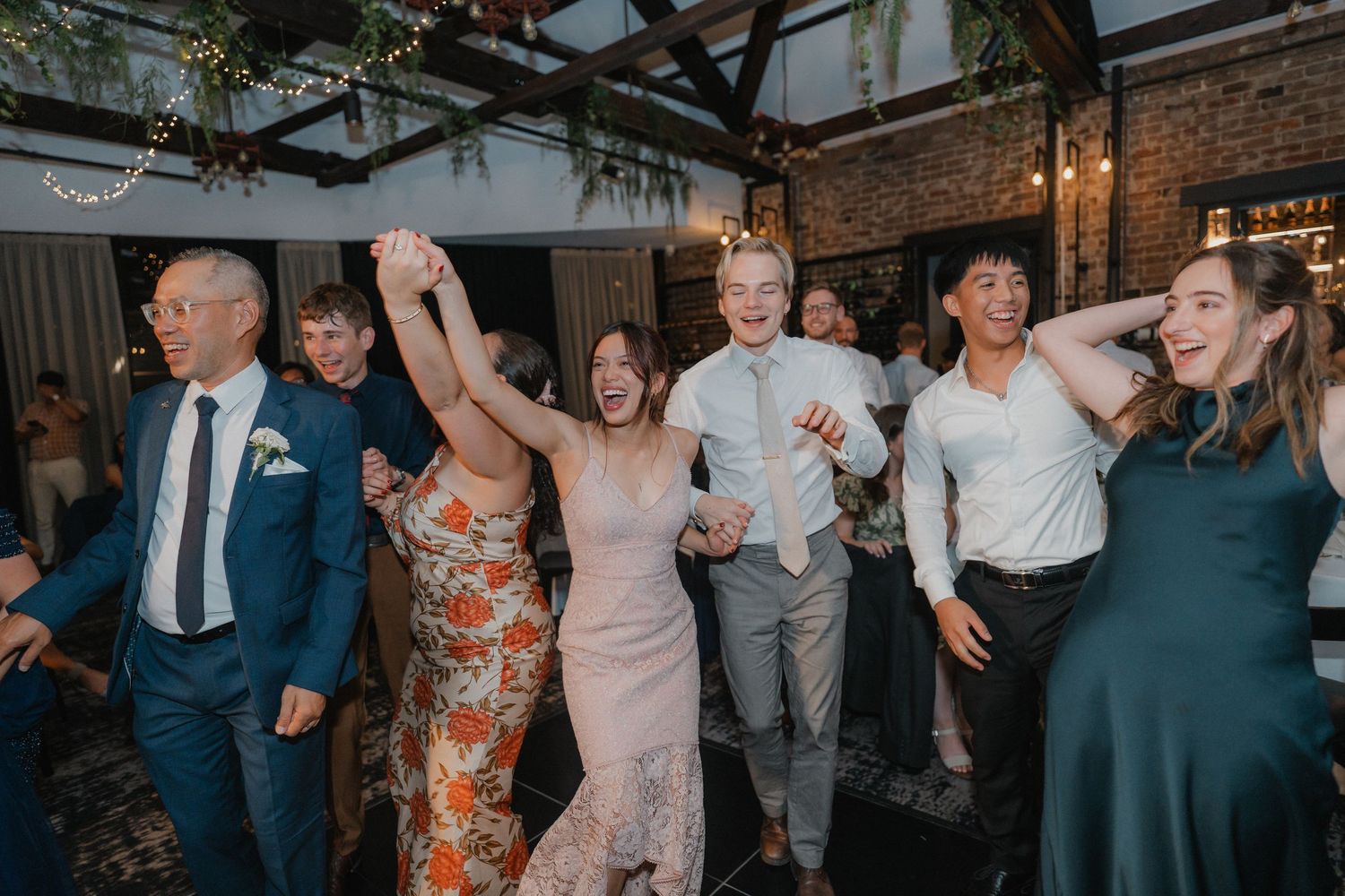Wedding guests dancing and celebrating energetically on a dance floor decorated with hanging lights.