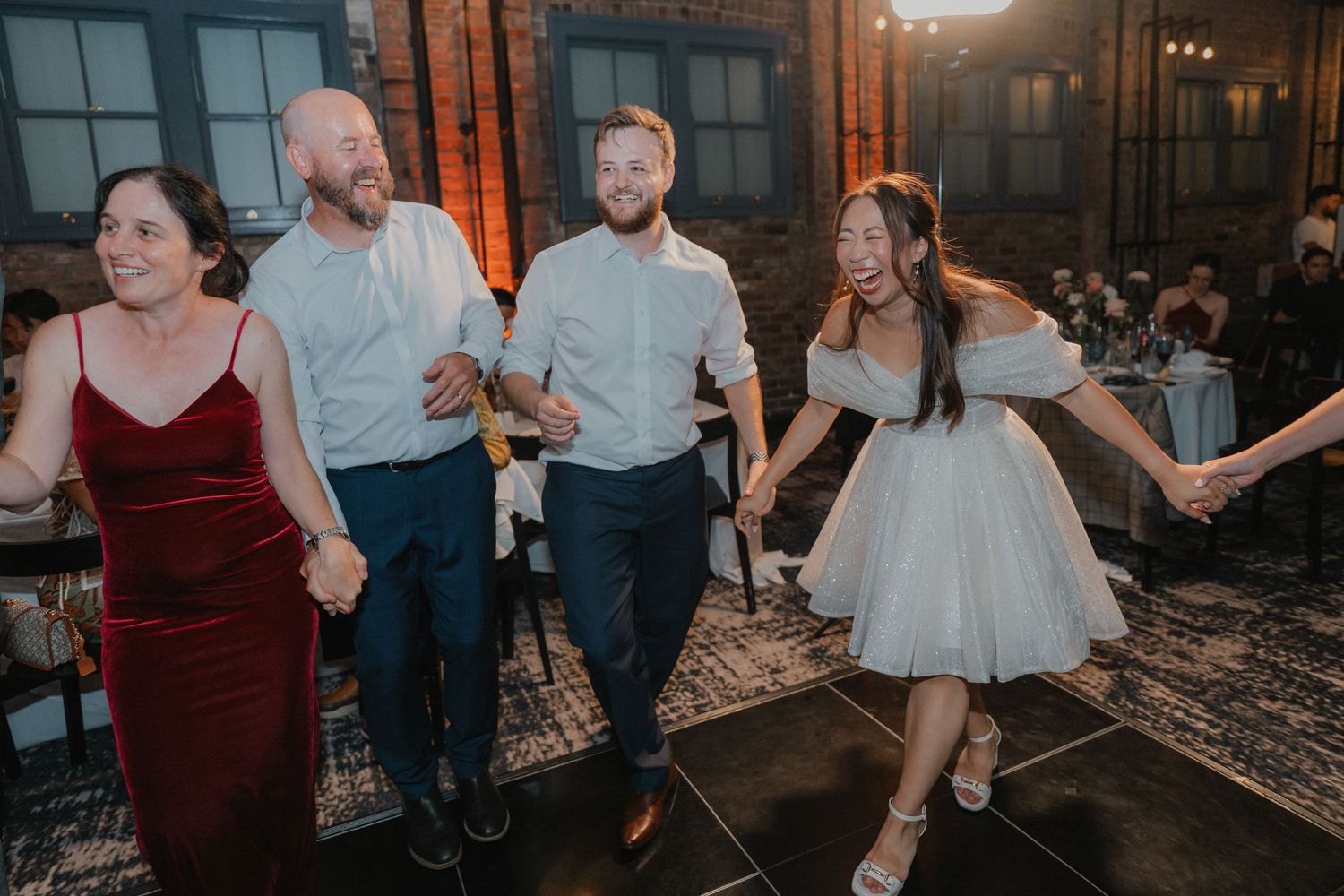 Wedding guests dancing in line at reception with exposed brick walls and vintage lighting.