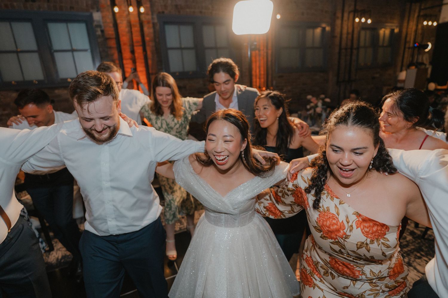 Wedding guests dance together joyfully at a dimly lit reception with warm lighting and festive atmosphere.