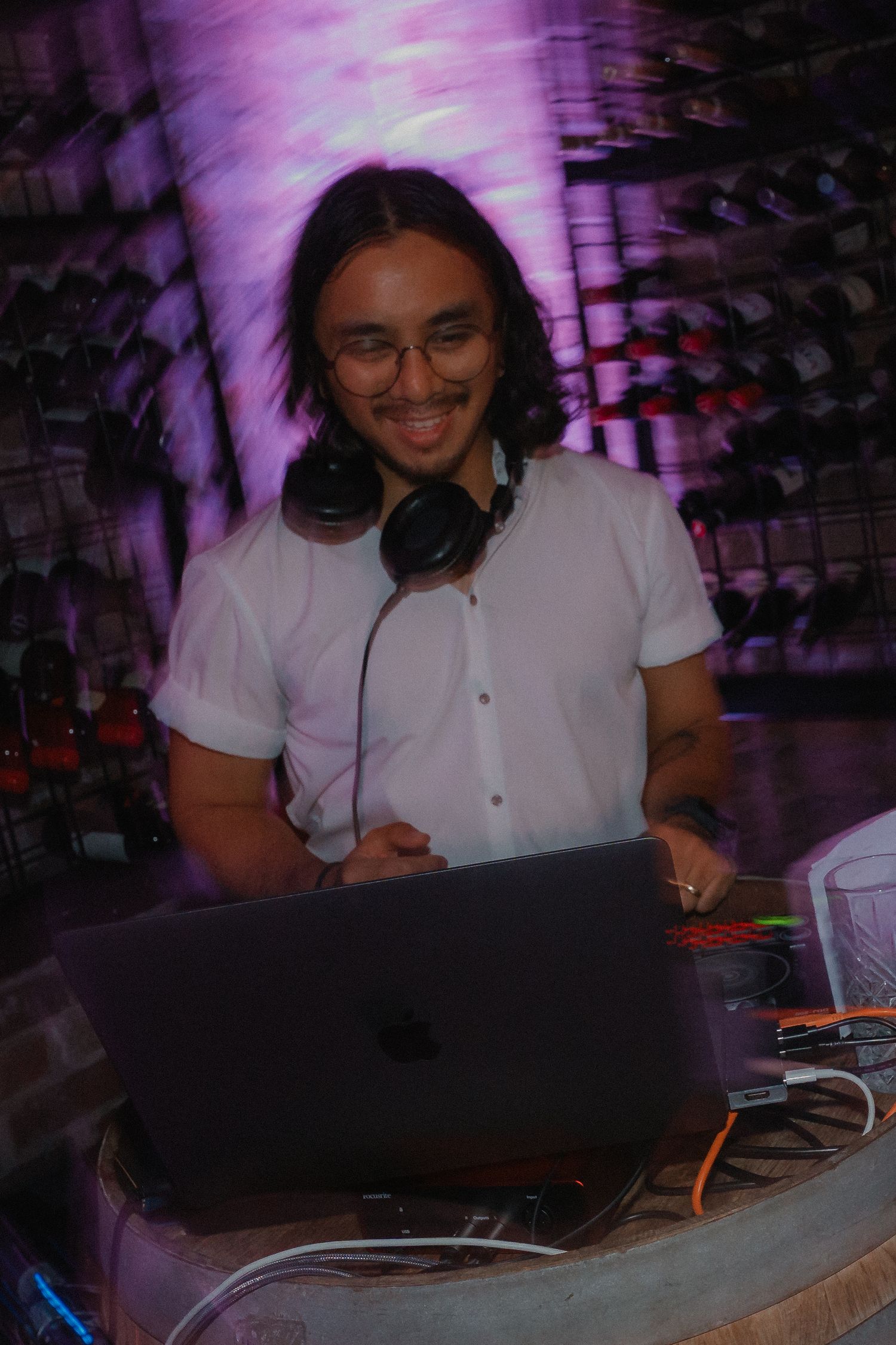 DJ working at a laptop with headphones in a purple-lit venue setting.