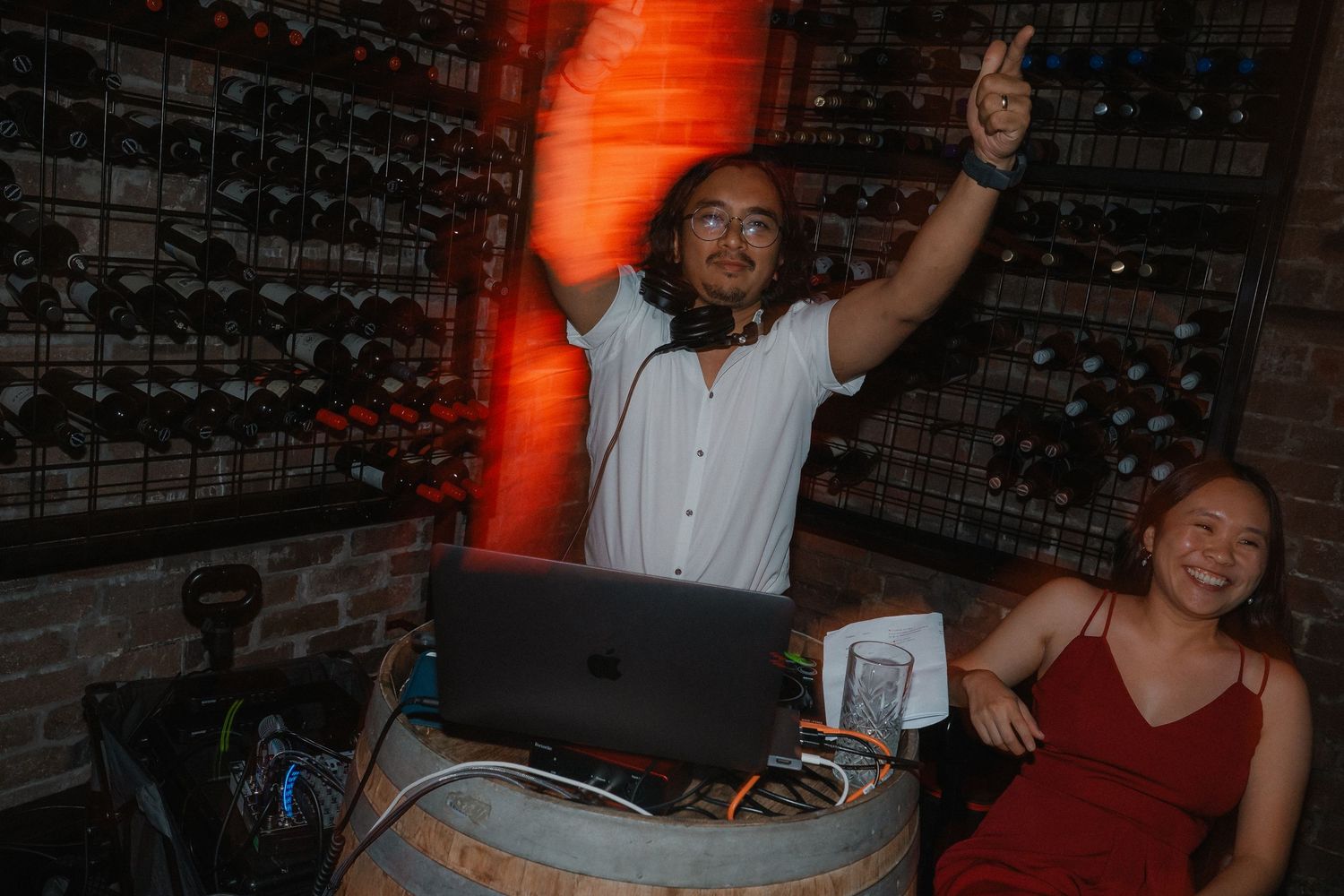 DJ performs behind laptop setup at nightclub with orange lighting and exposed brick wall in background.