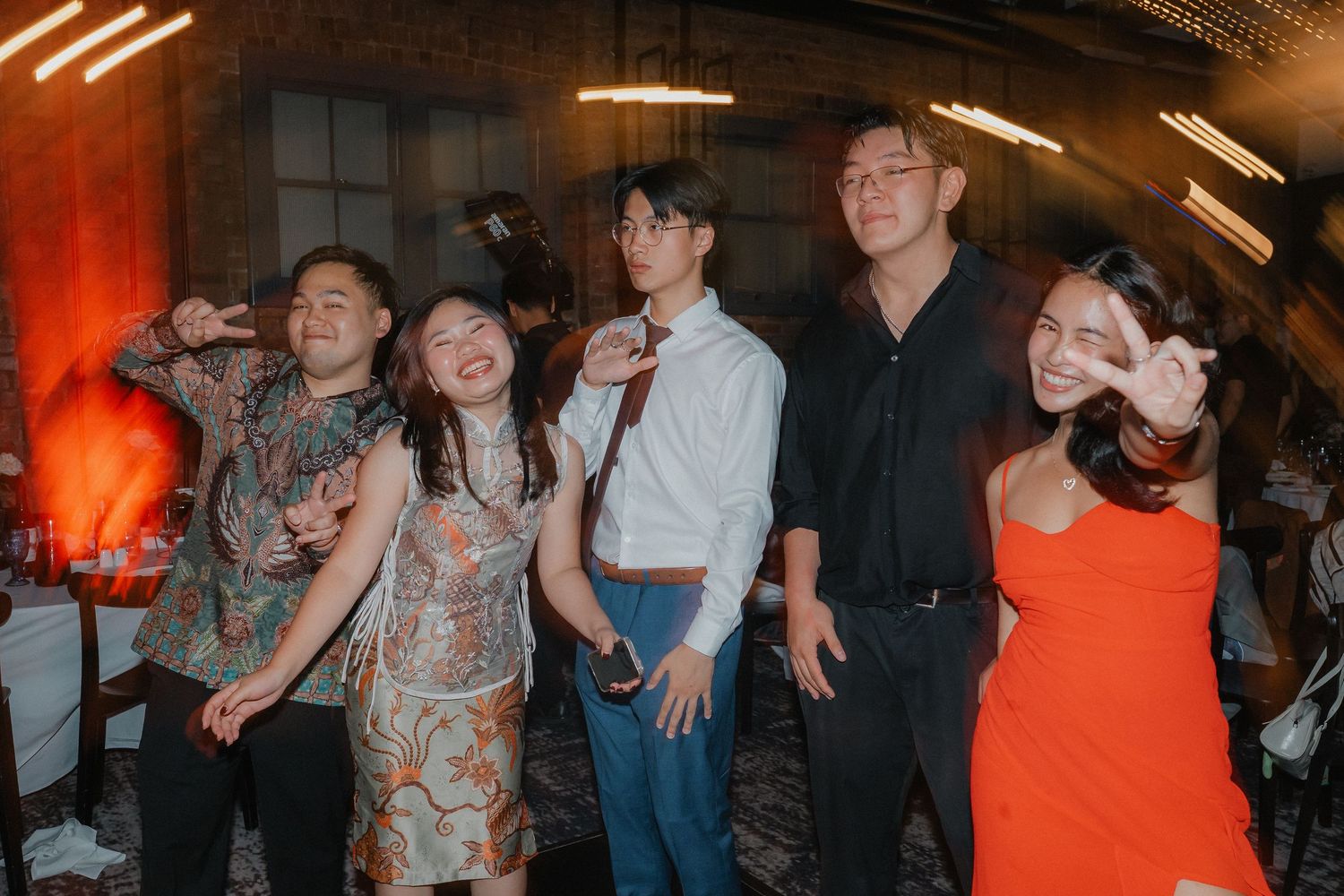 Wedding party celebrating with energetic dancing and group hugs at a reception with warm red lighting.