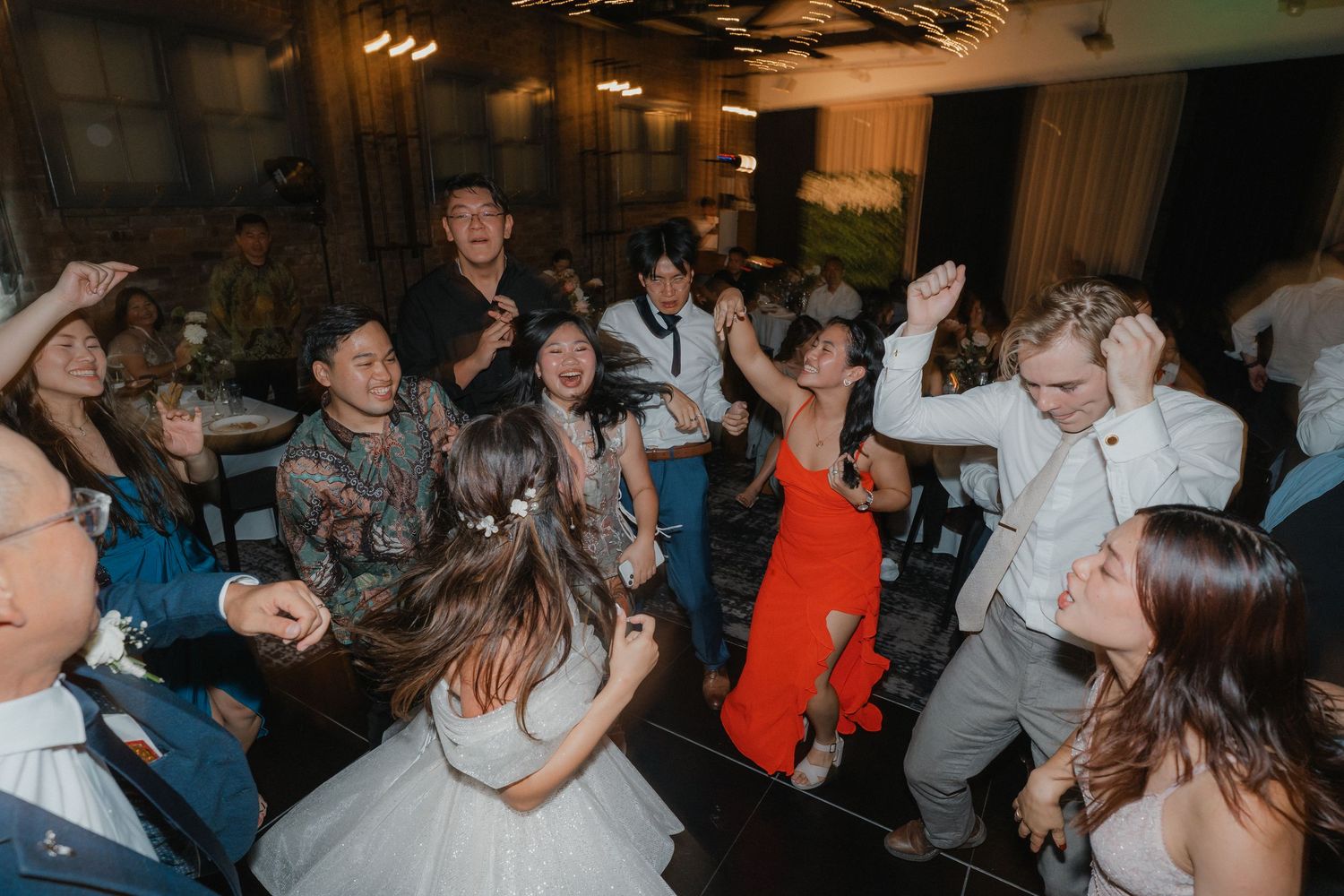 Wedding guests enjoying themselves on a lively dance floor at an indoor reception celebration.