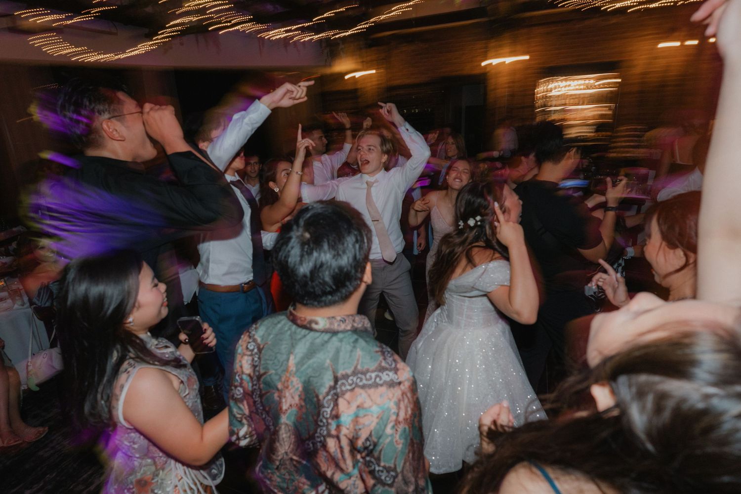Celebratory crowd dances in a packed venue under moody lighting at a wedding reception.