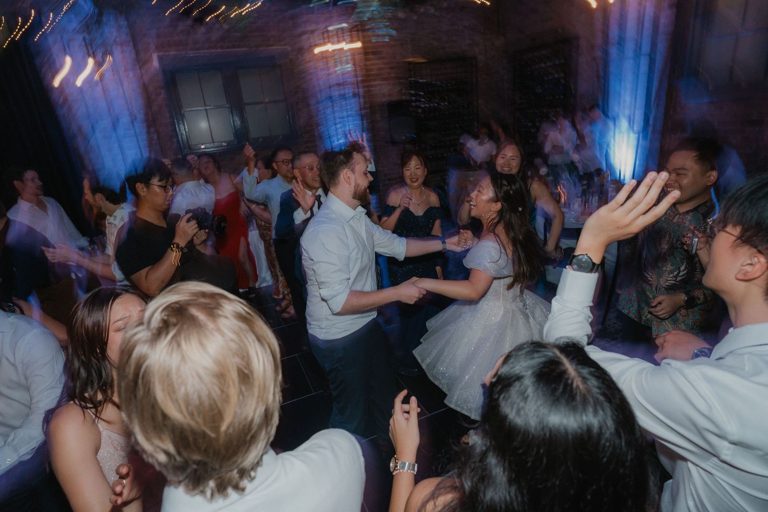 A large group of wedding guests dance energetically at a dimly lit reception venue with blue lighting.
