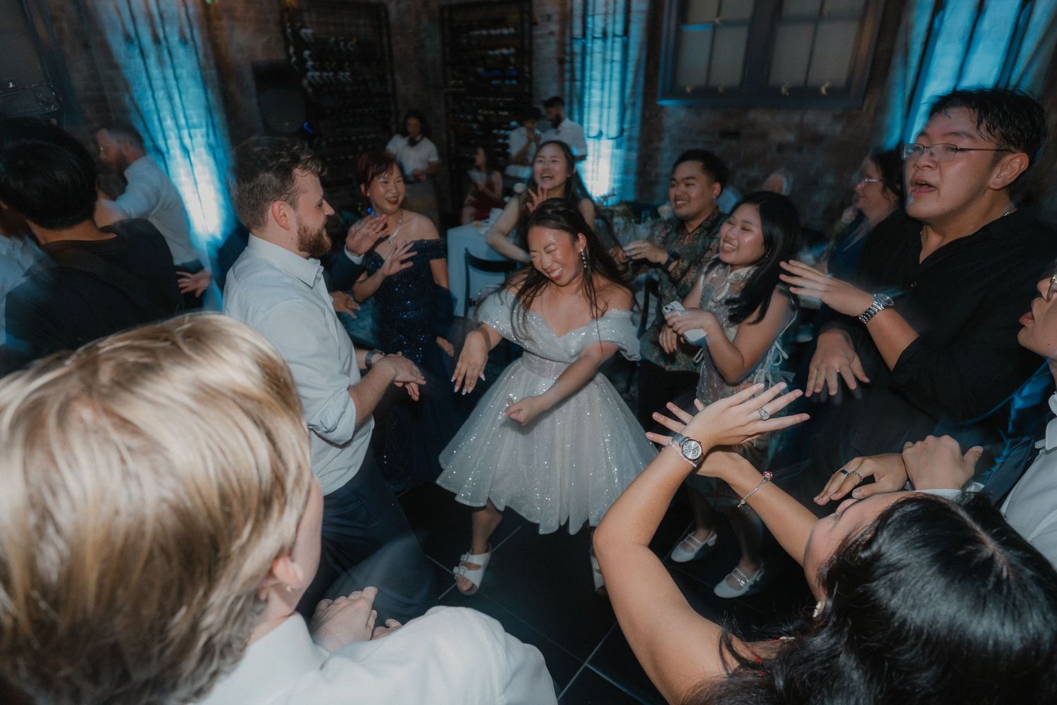 Wedding party forms an energetic circle on the dance floor under blue accent lighting.