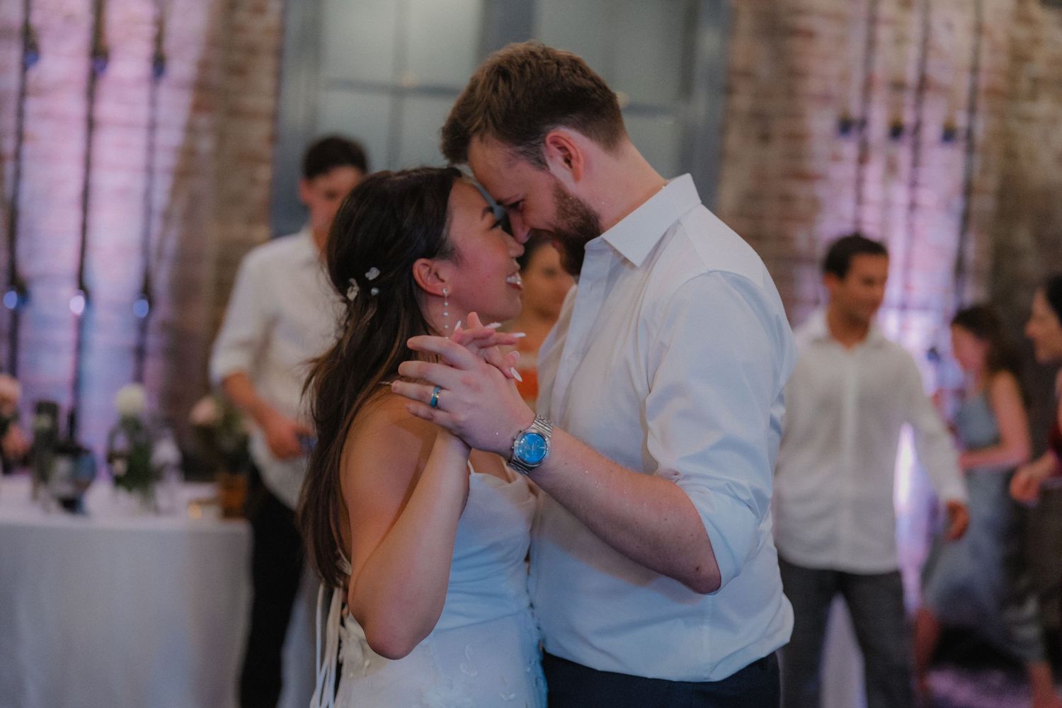 Couple sharing an intimate dance moment in a rustic brick-walled reception venue with purple lighting.