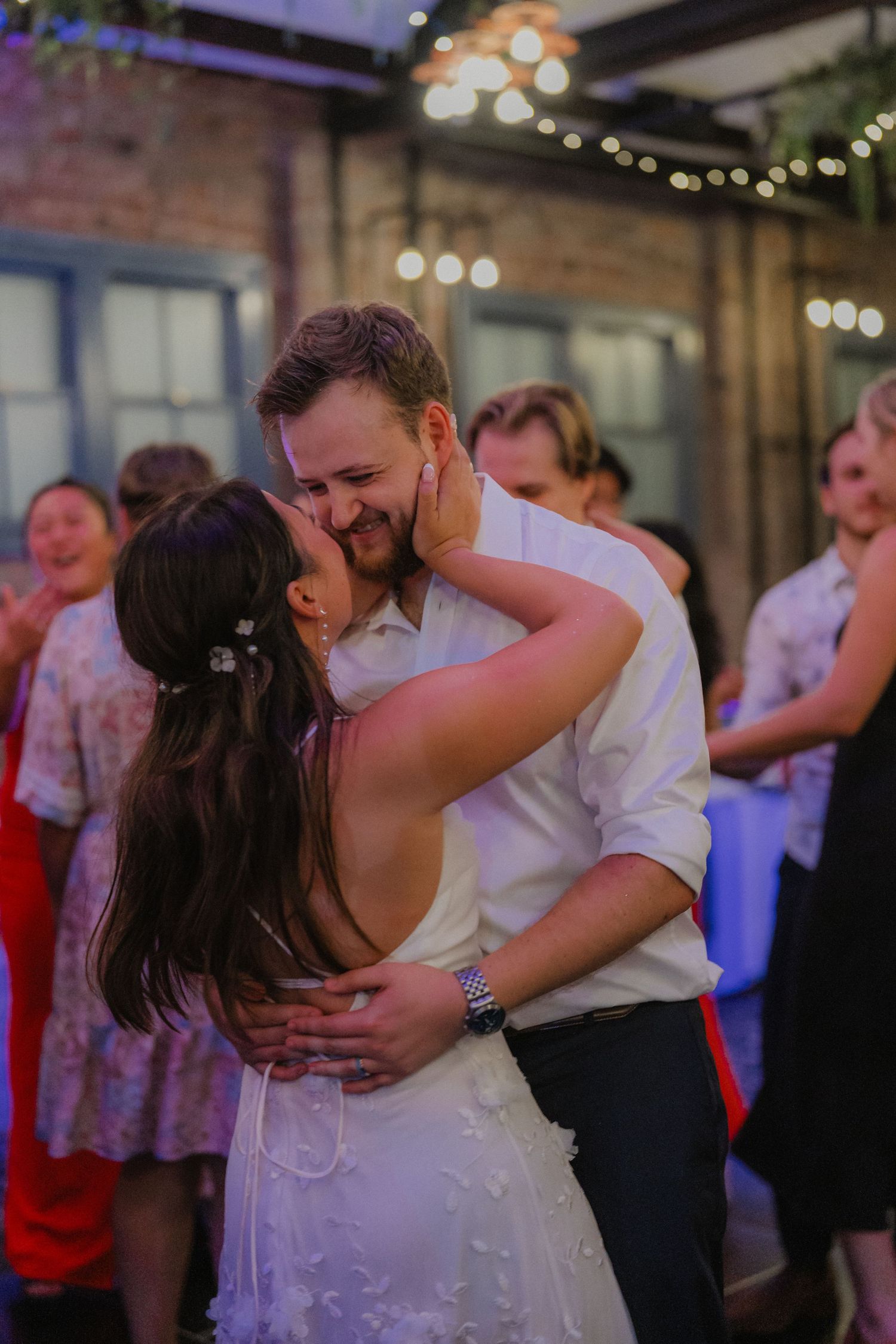 Newlywed couple shares a romantic first dance in an industrial-style venue with exposed brick and string lights.