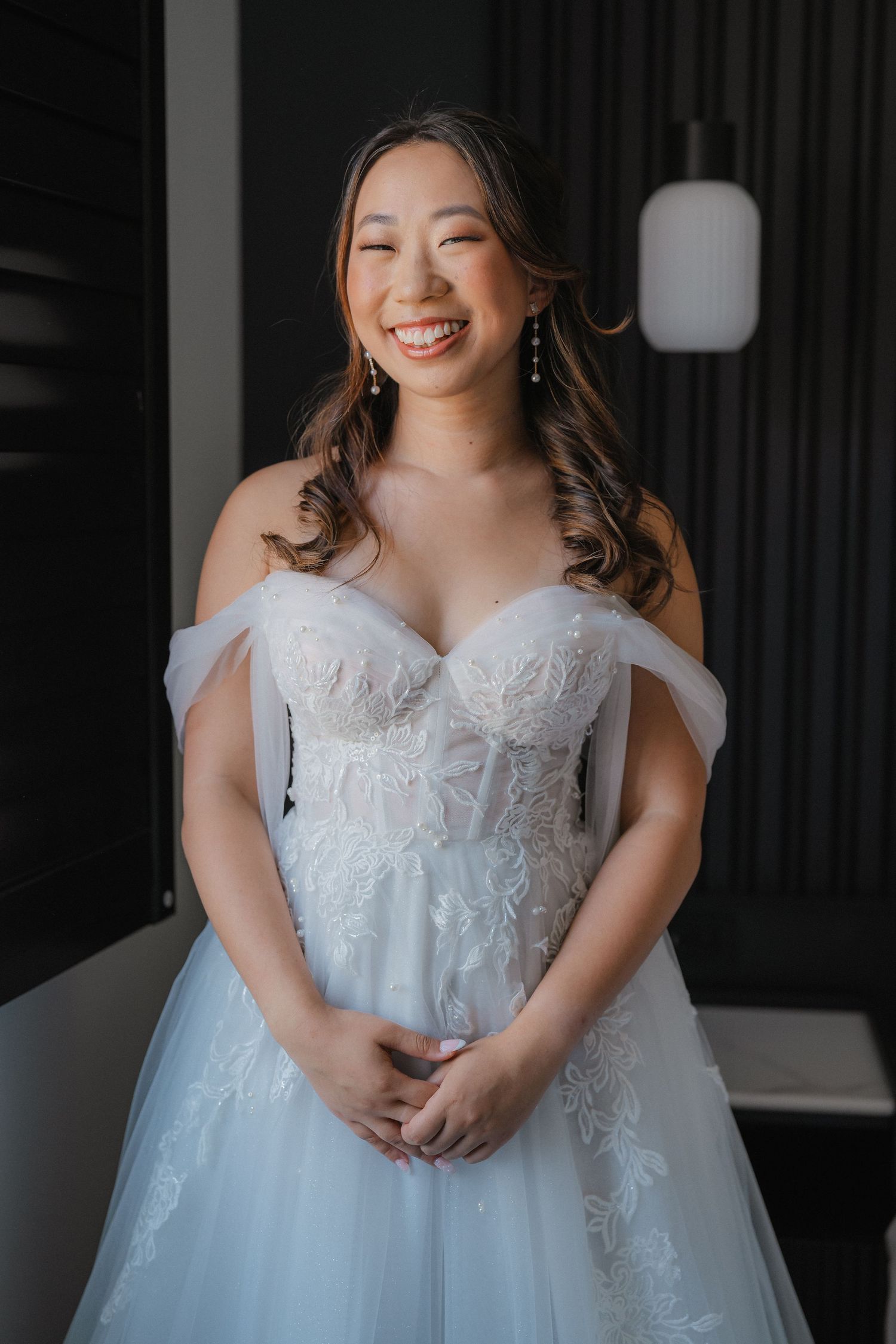 A beautiful bride poses in an elegant white lace wedding gown with a flowing tulle skirt in an indoor setting.