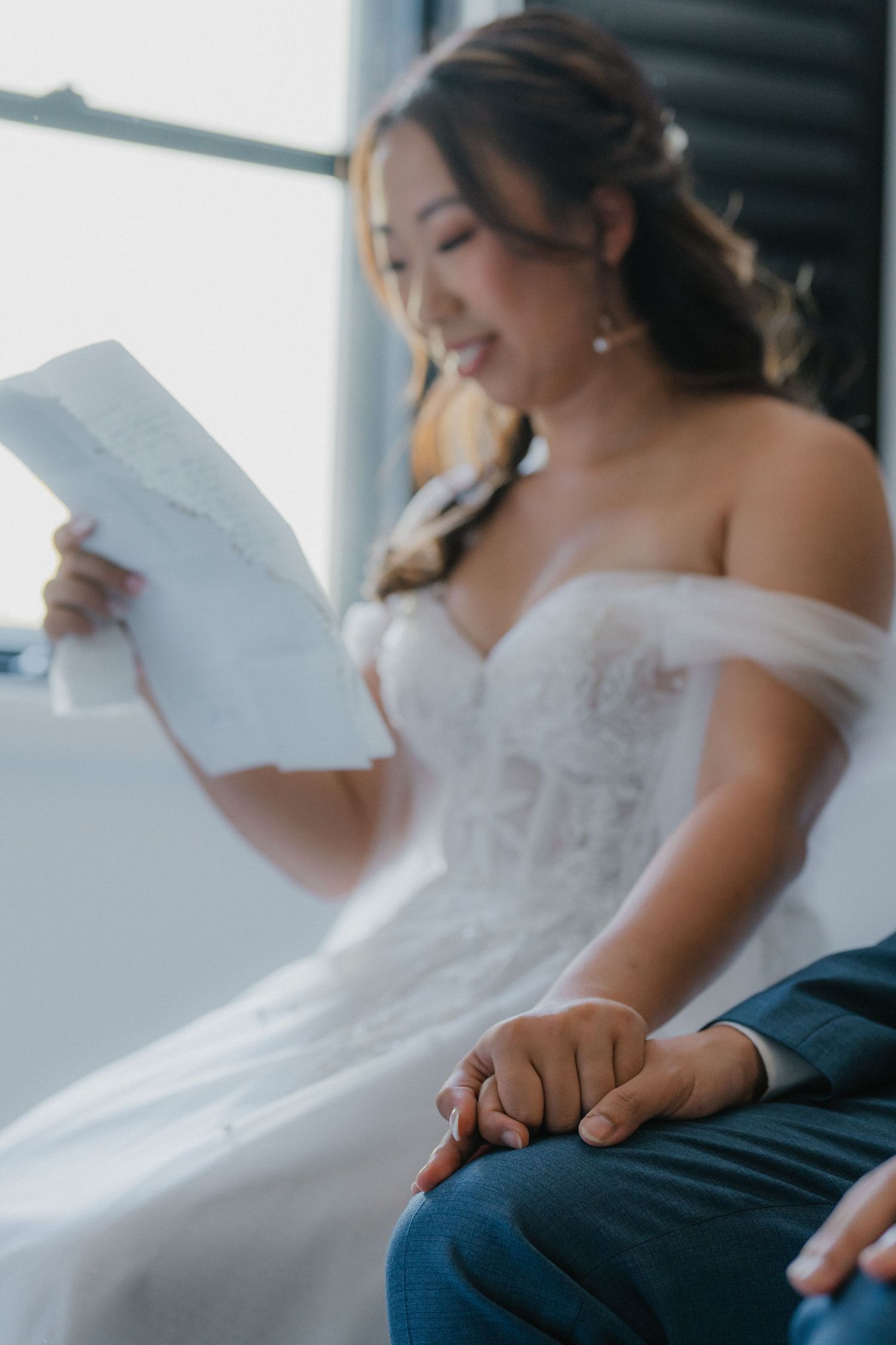 Bride in an off-shoulder wedding dress reading a letter while seated.
