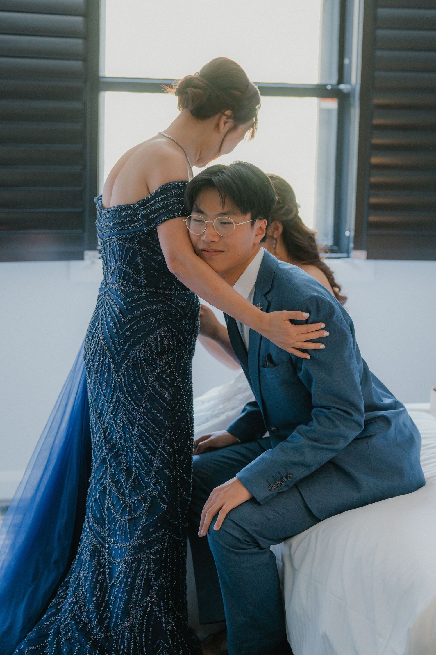 Two people in formal attire embracing each other on a white couch near a window with shutters.