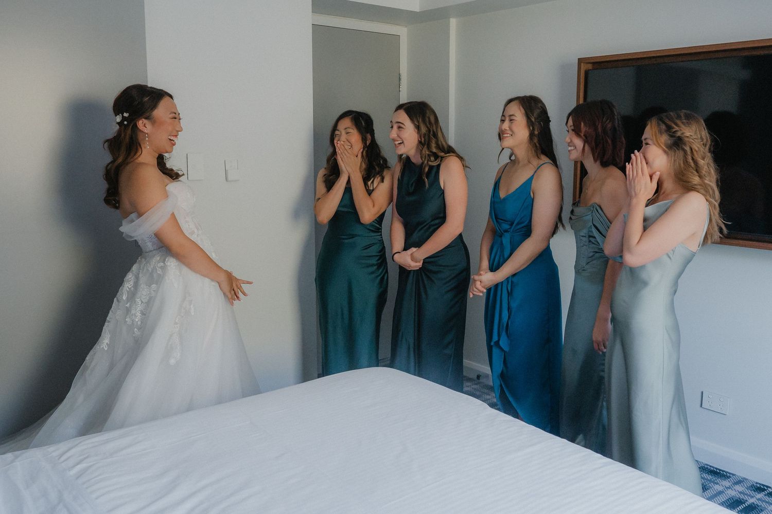 A bride in white shows off her dress to bridesmaids in blue dresses who react with excitement.