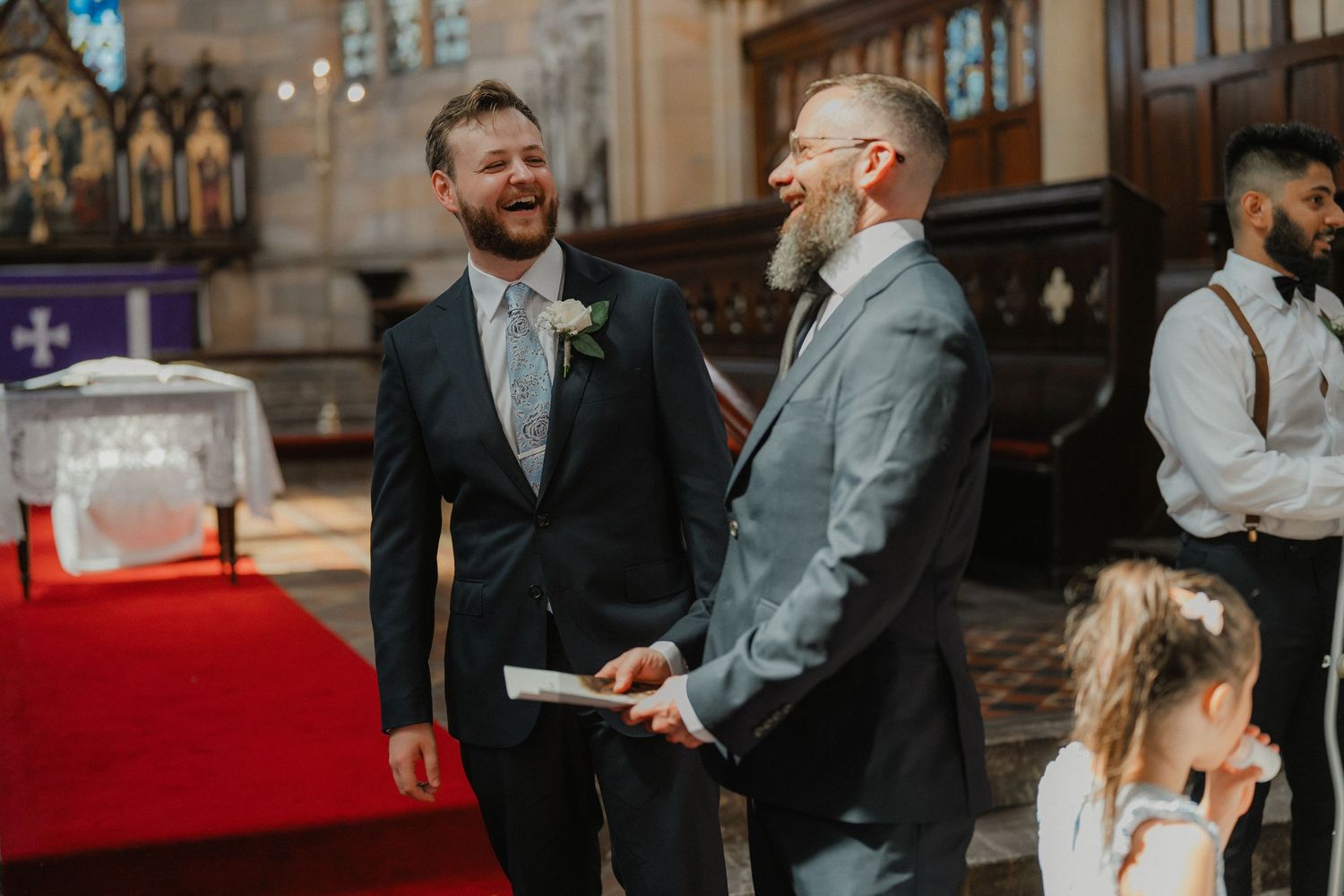 Two wedding attendees share a candid conversation inside a grand church with gothic architecture and red carpet.