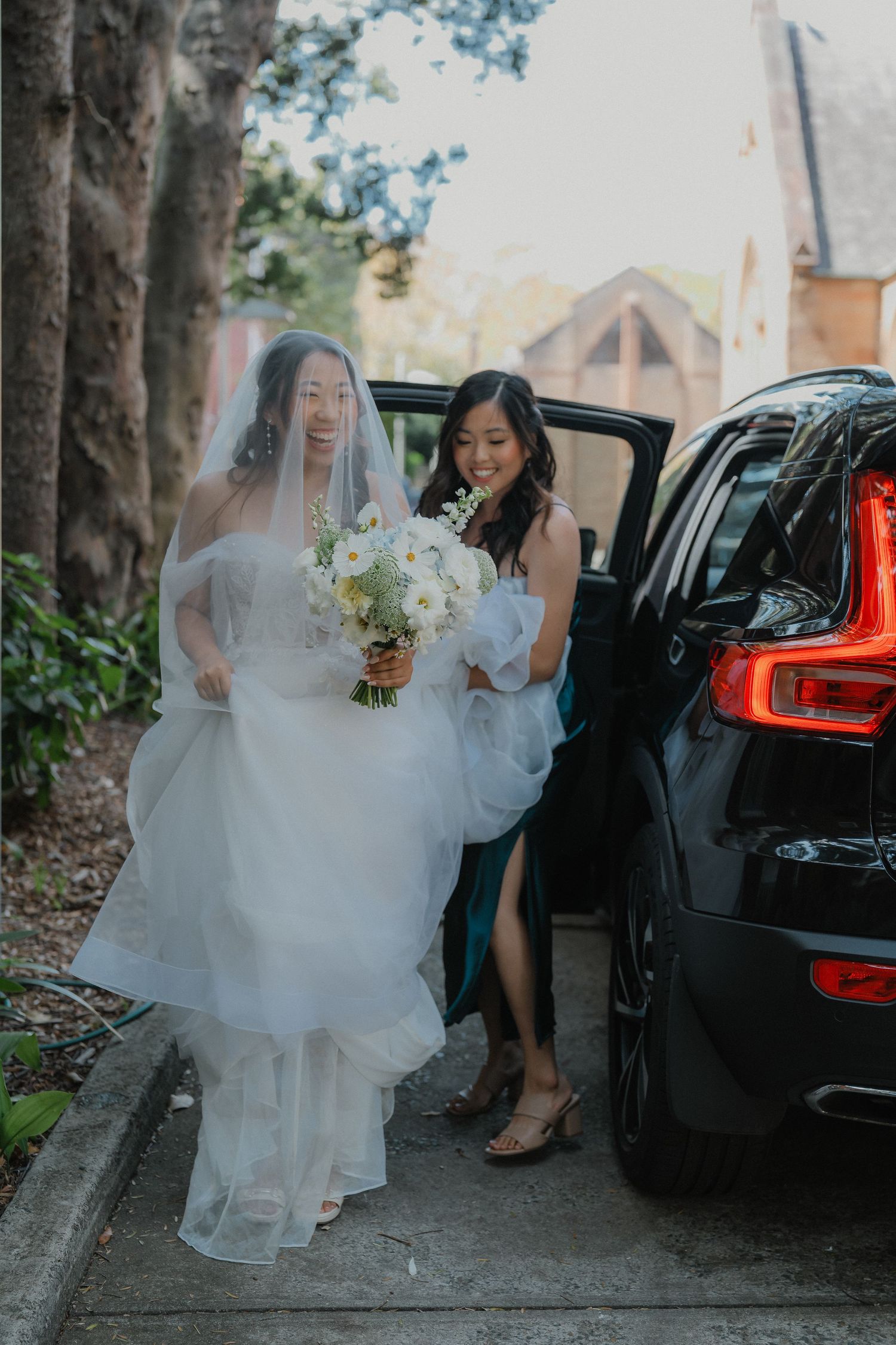 A bride in flowing white dress exits a black car while holding a white floral bouquet.