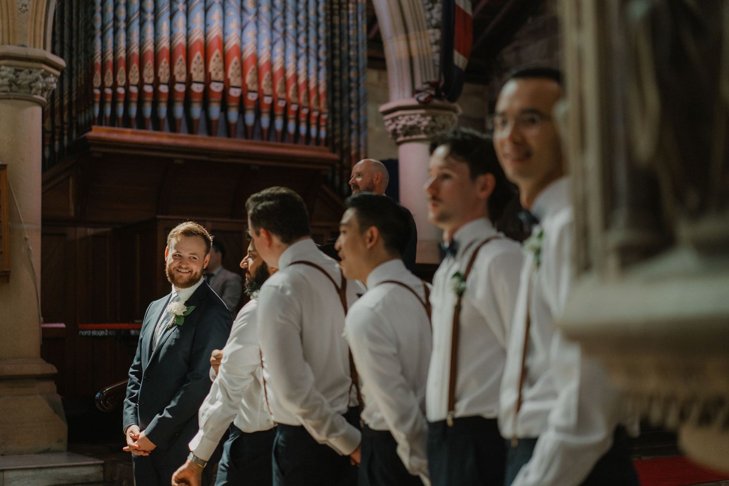 Groomsmen stand in formation inside an ornate church with pipe organs and stone columns.