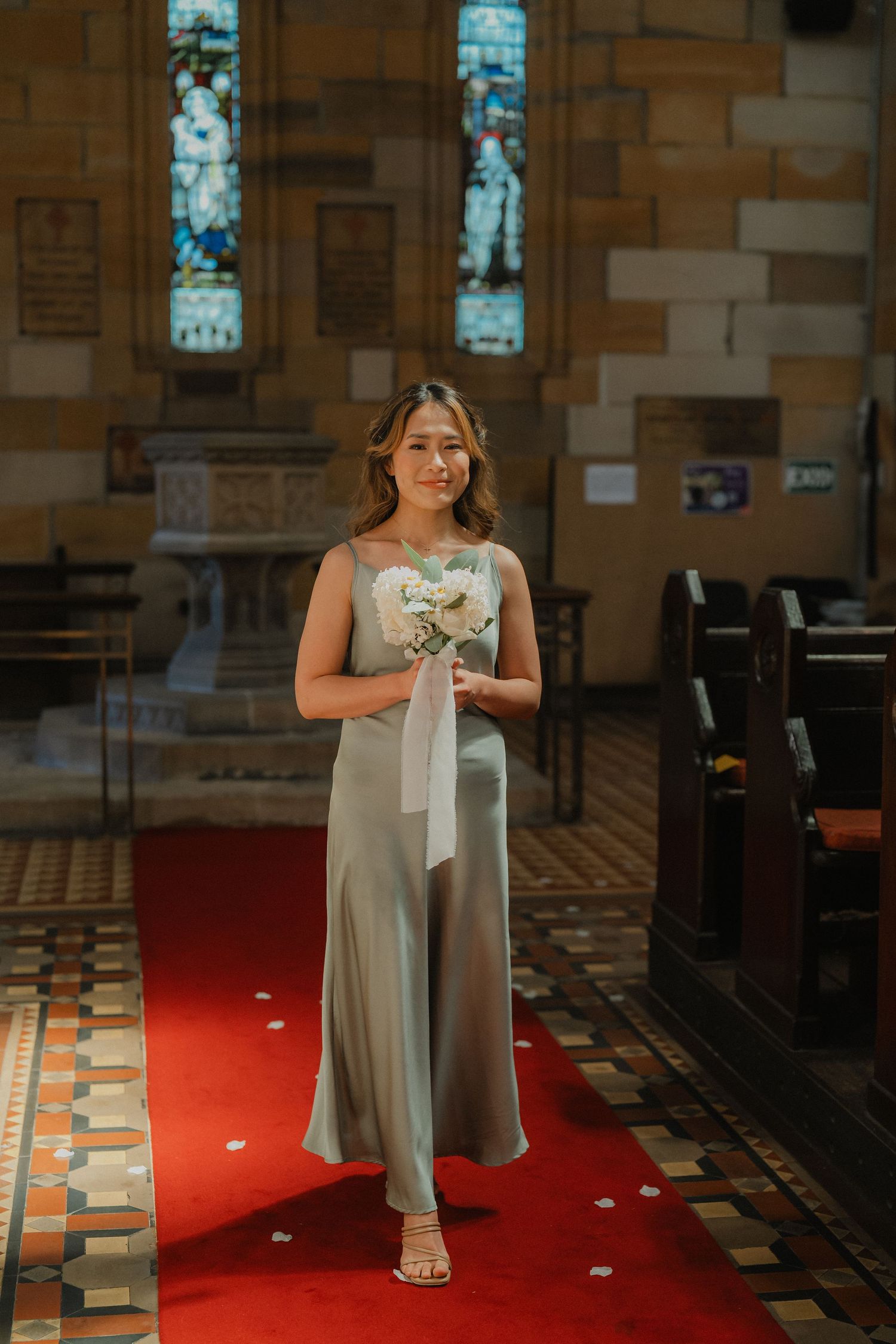 A bridesmaid walks down a red-carpeted church aisle wearing a flowing sage green dress and holding a white bouquet.