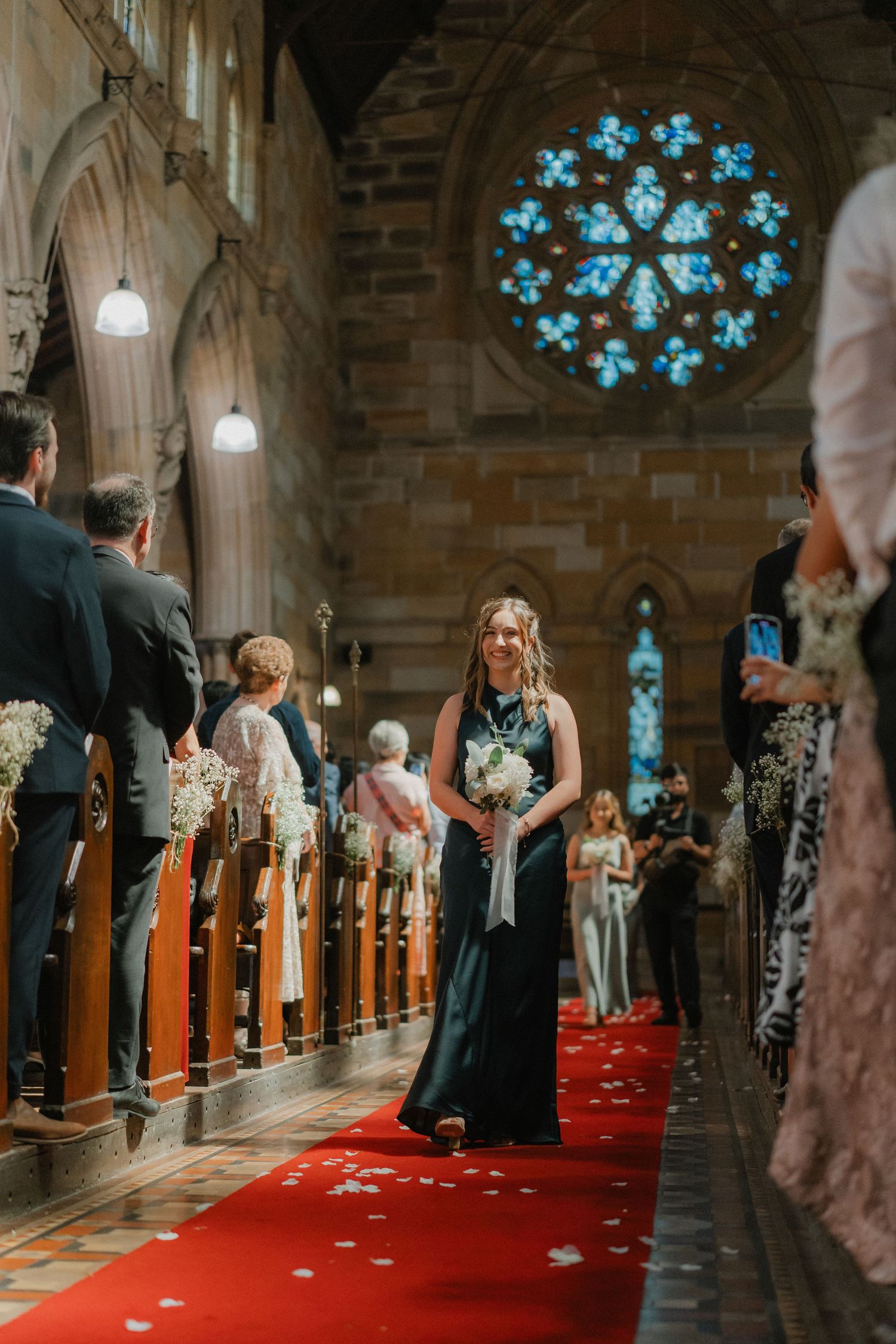A church wedding procession showing stained glass windows and guests along wooden pews on a red carpet.