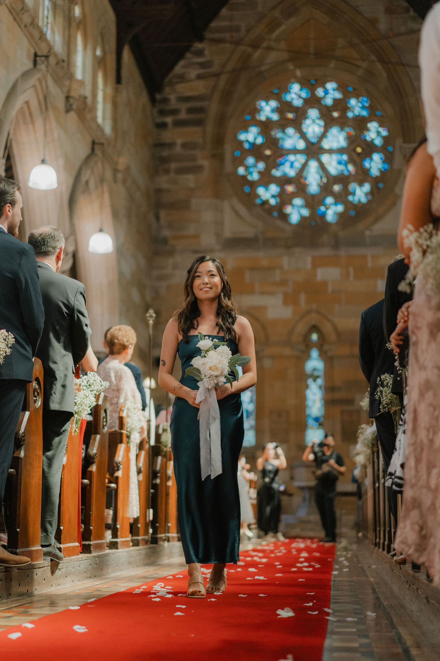 A bridesmaid walks down the red-carpeted church aisle beneath a stained glass window during a wedding ceremony.