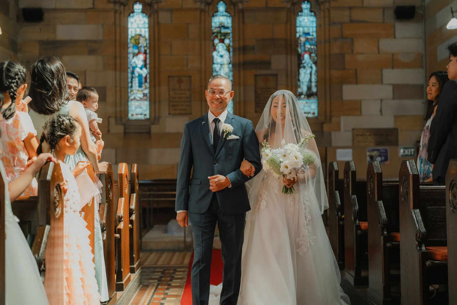 A wedding ceremony processional takes place in a beautiful historic church with stained glass windows.