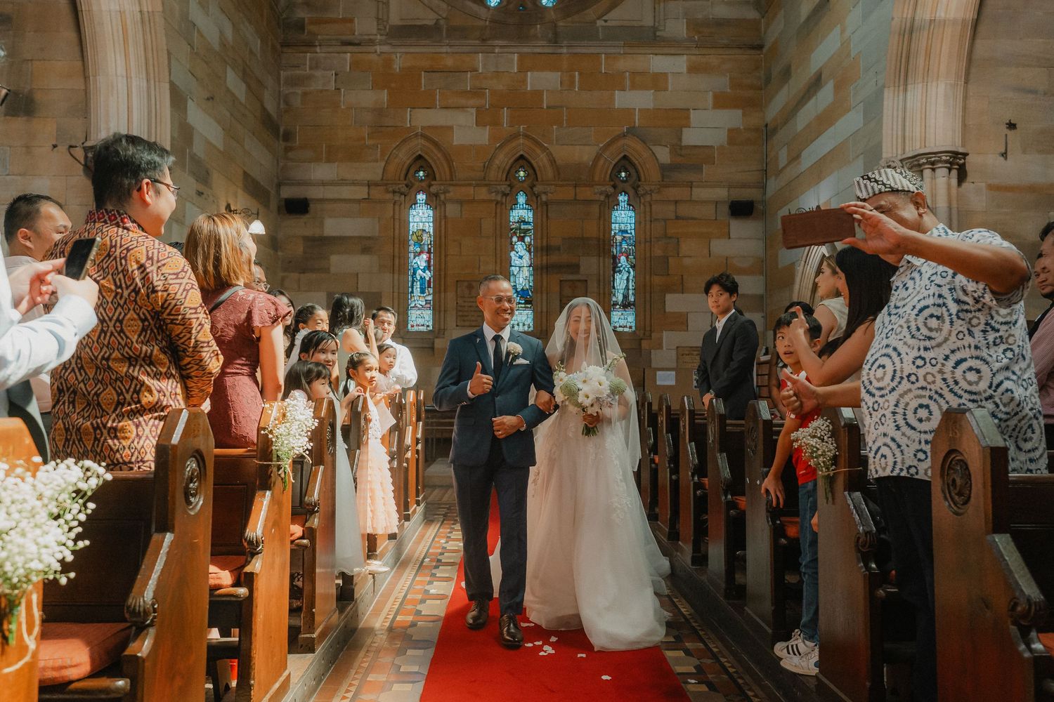 Wedding ceremony in progress with guests standing in ornate church interior with red carpet.