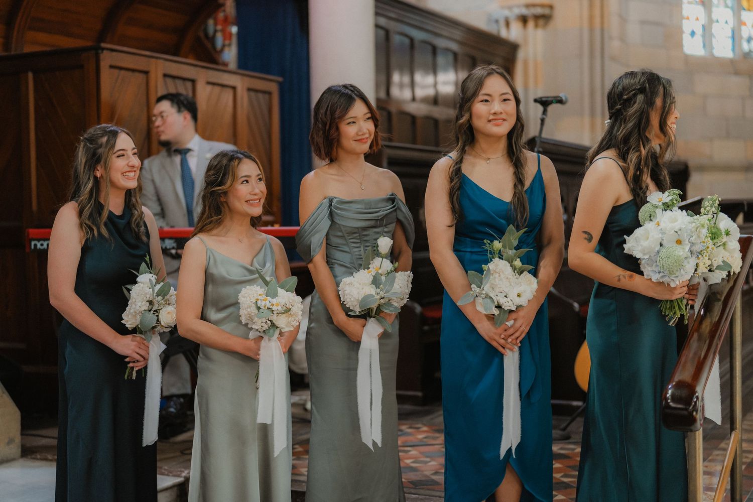 Bridesmaids in elegant teal and grey dresses hold white bouquets during a church wedding ceremony.