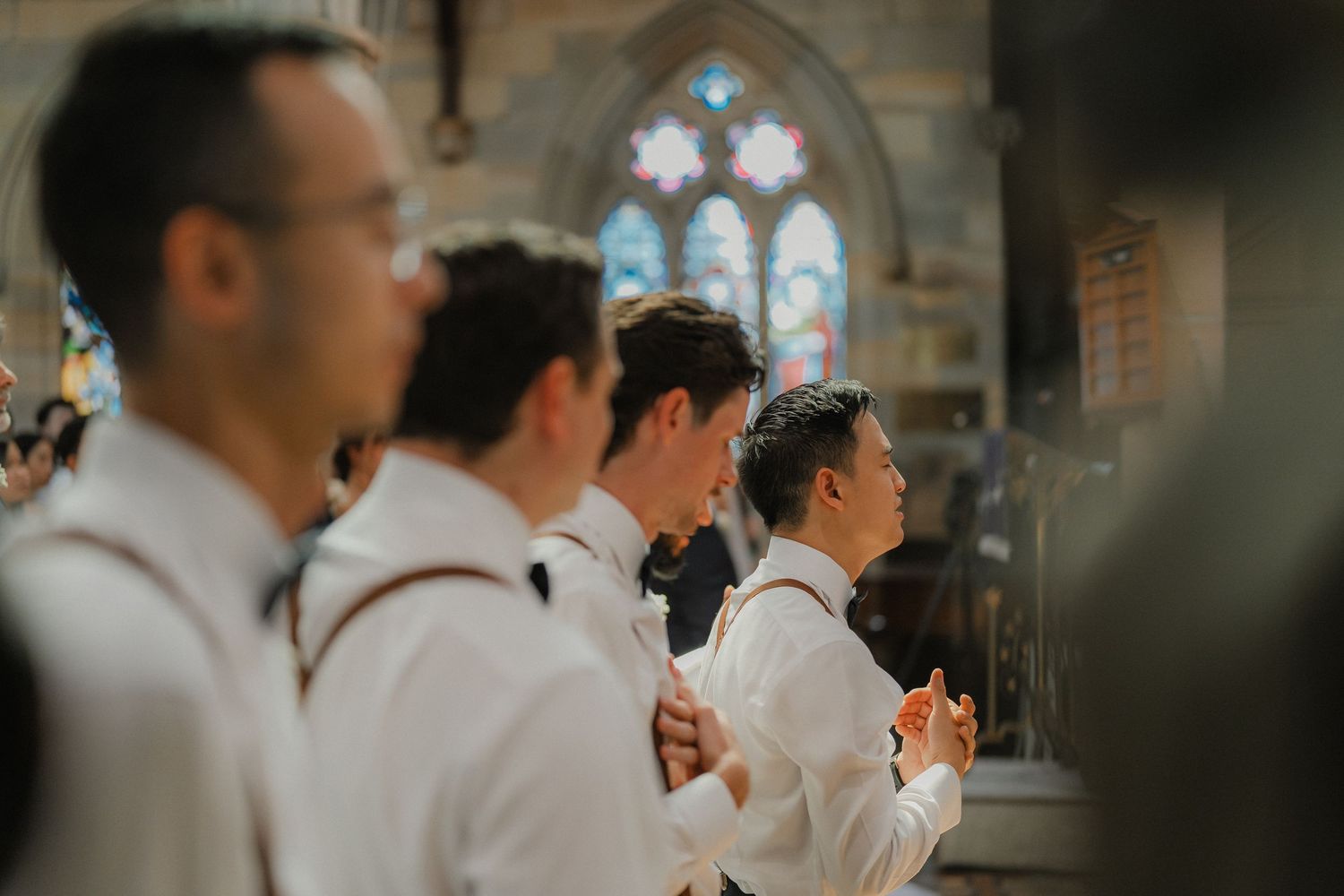 Groomsmen in white dress shirts and suspenders stand in formation at a church ceremony.