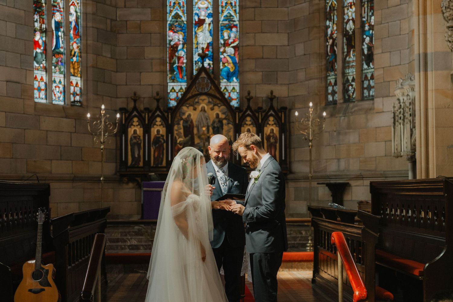 Wedding ceremony takes place at the altar of a historic stone church with stained glass windows and gothic architecture.