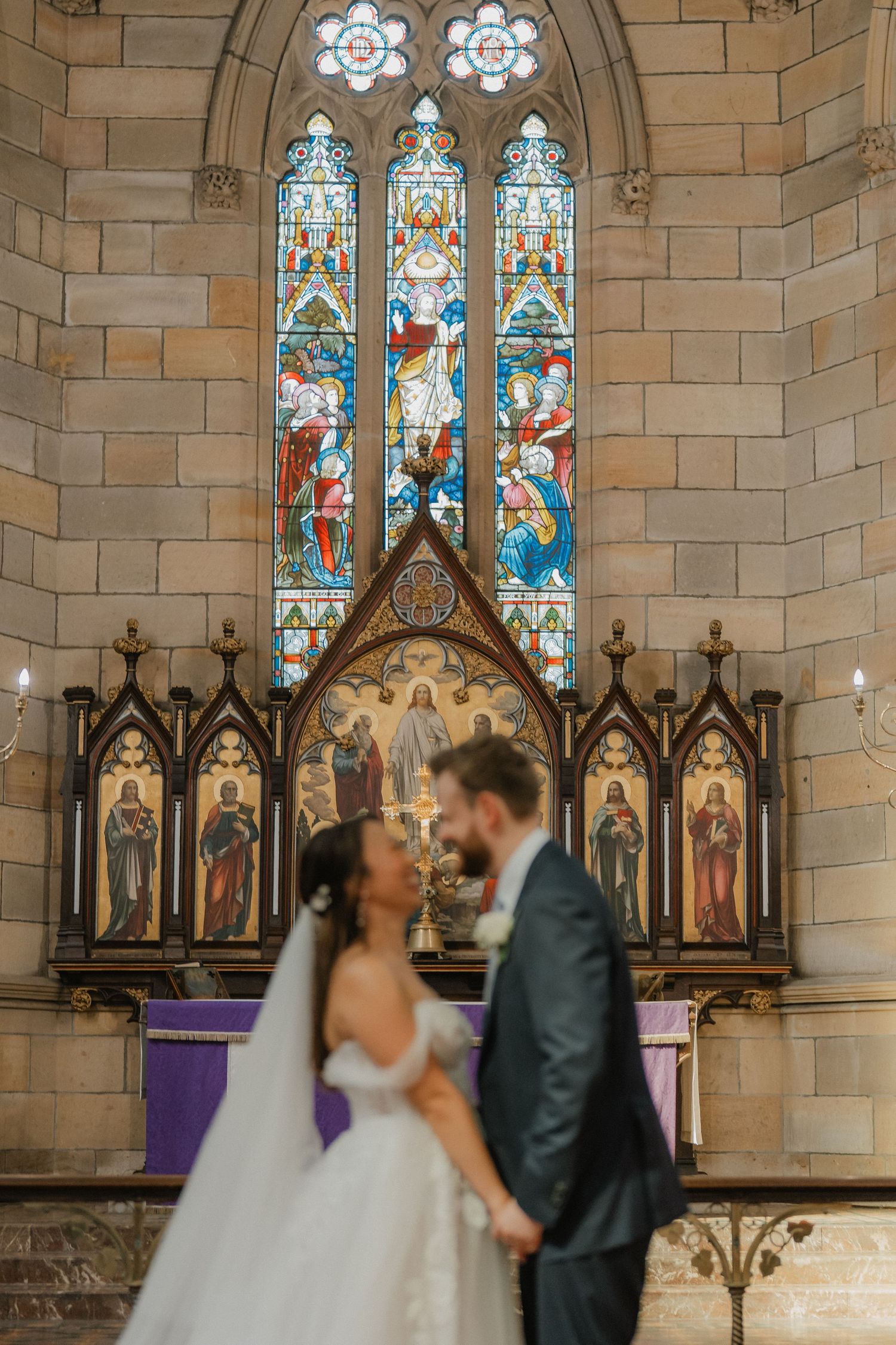 A wedding couple shares an intimate moment in front of stained glass windows and ornate church altar.