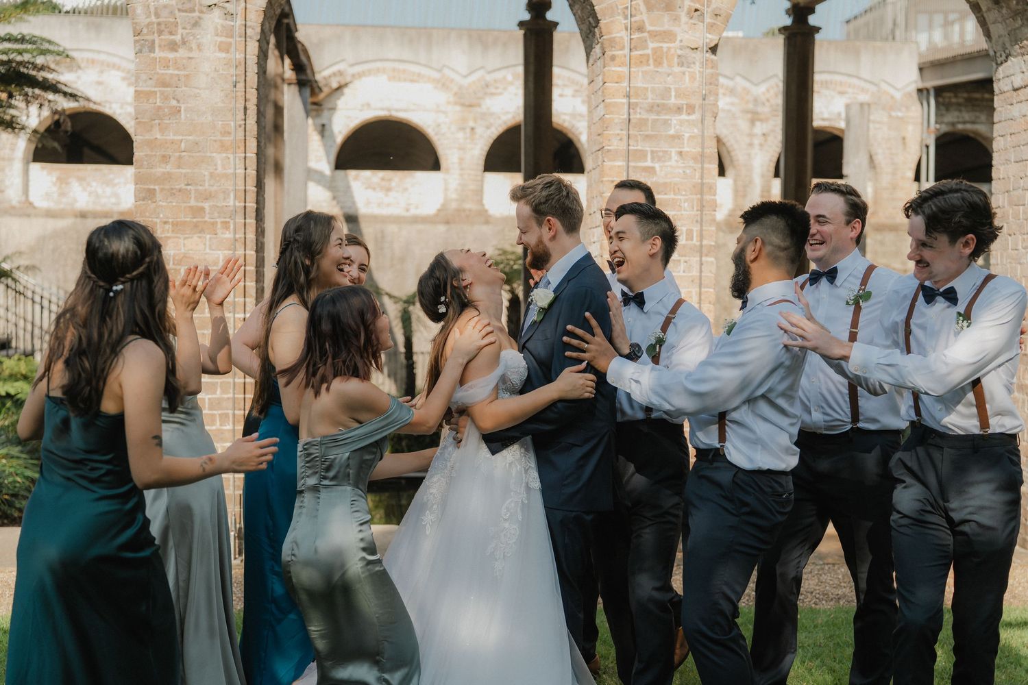 Wedding party celebrates outdoors with bridesmaids in green and groomsmen in light blue against stone architecture.