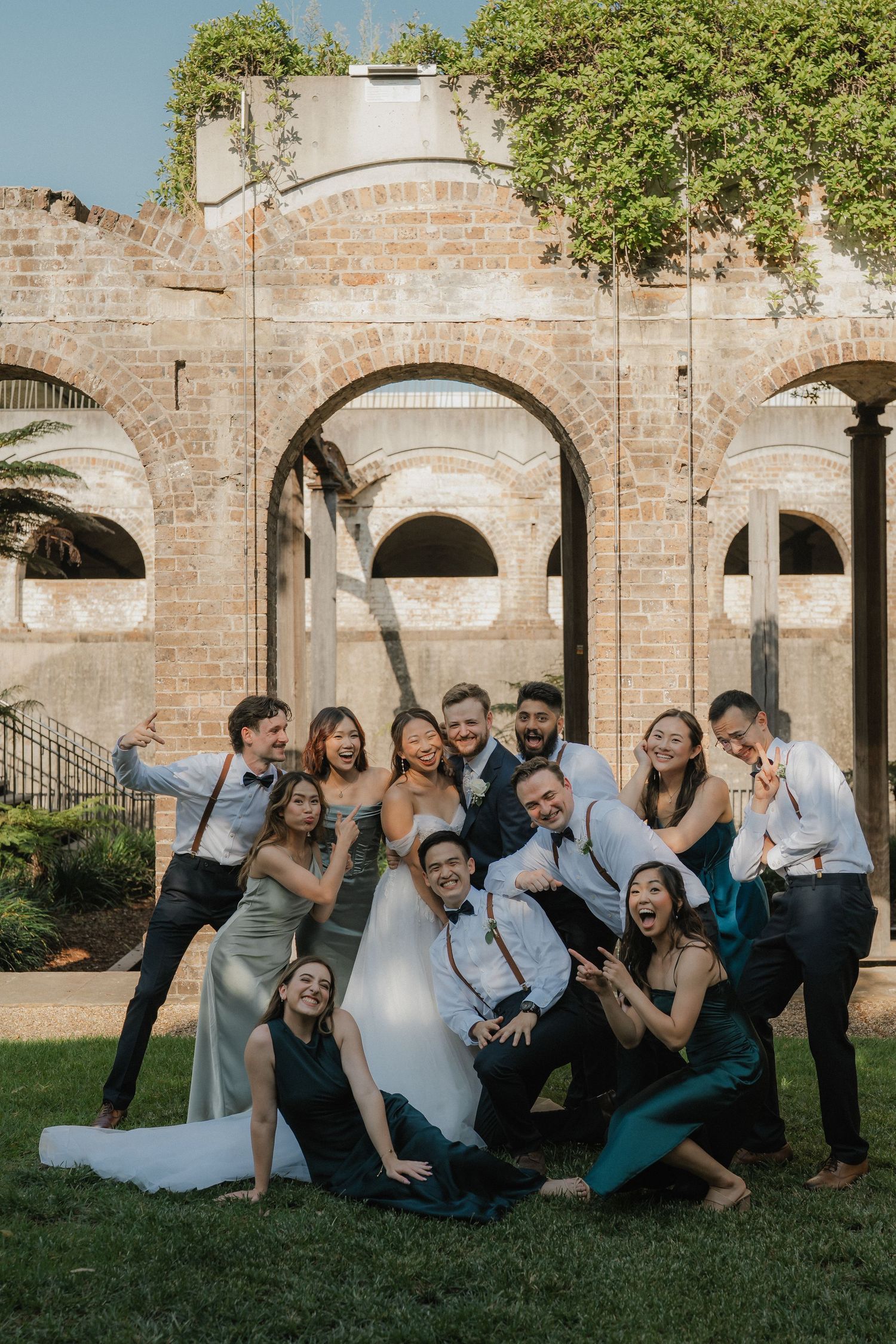 Group poses playfully on grass in front of rustic brick archways during outdoor wedding photo session.