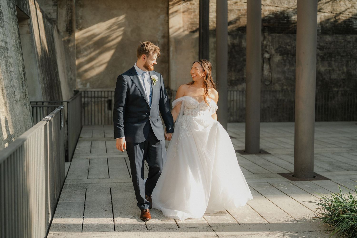 A bride and groom walk together on a covered walkway bathed in sunlight and shadows.
