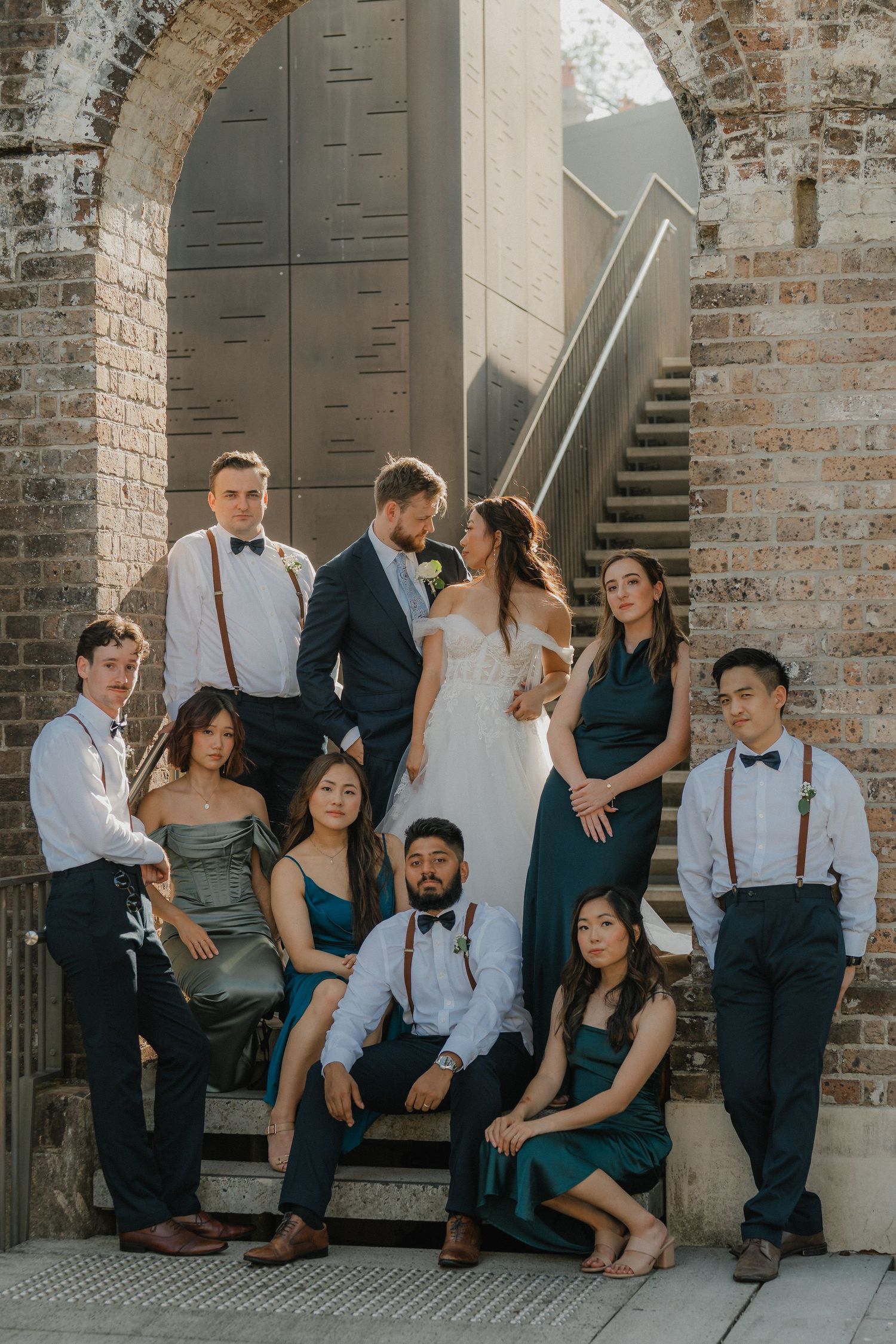 Wedding party poses on brick stairs wearing elegant attire in navy and white with suspenders and bow ties.