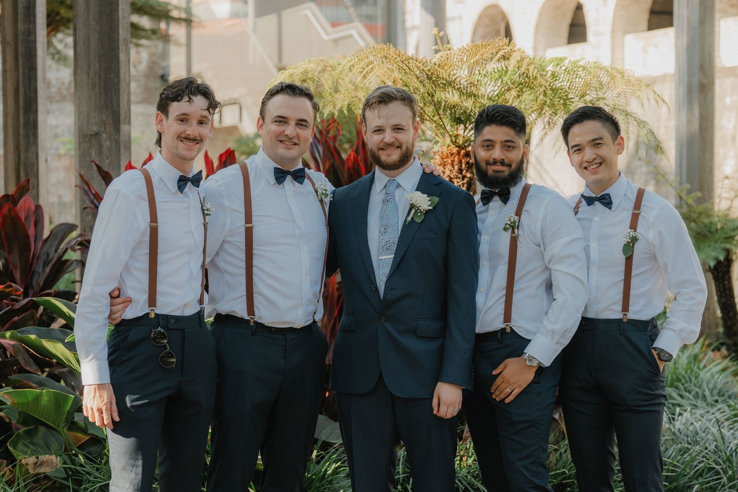 Groomsmen in suspenders and bow ties pose together outdoors for formal wedding party photos.