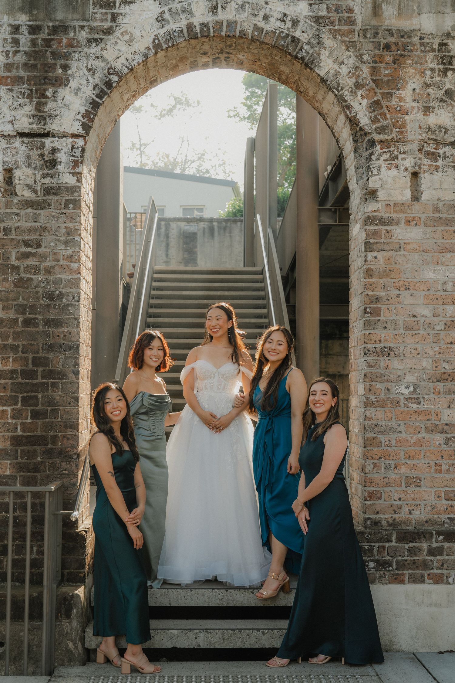 Bridesmaids in teal dresses pose with bride in white gown against rustic brick wall with arched windows.