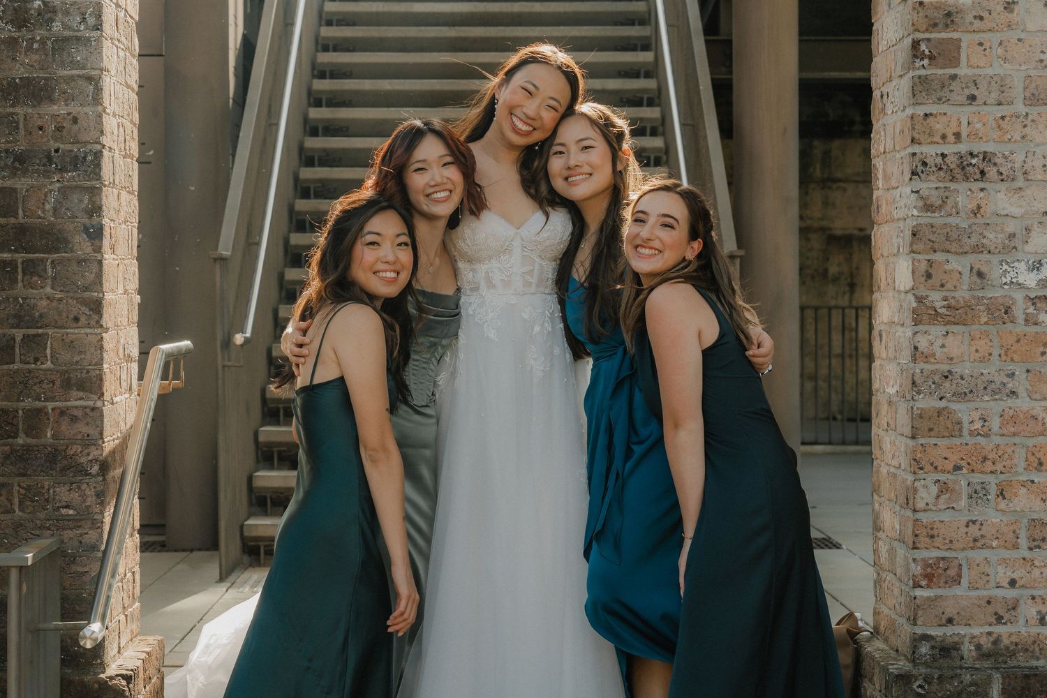 Wedding party in teal gowns gather close on brick staircase for photo.