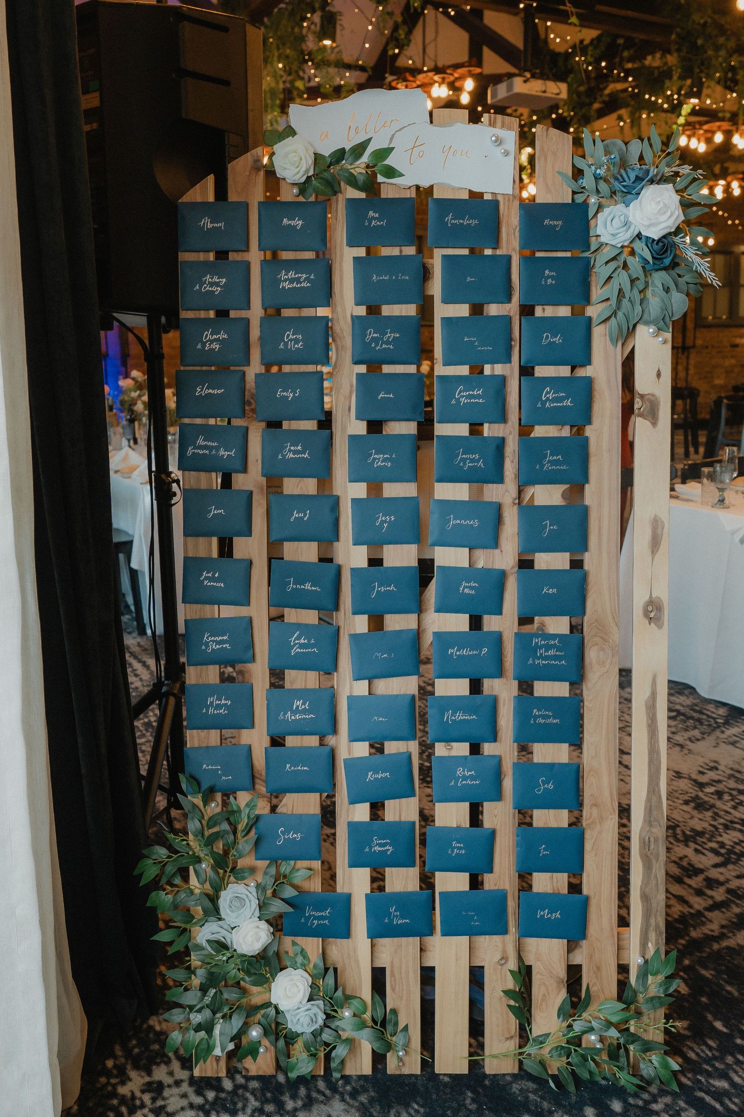 Blue wooden seating chart display decorated with greenery and string lights at wedding reception.