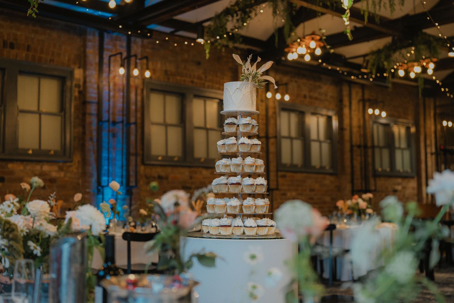 Elegant wedding reception venue features rustic wooden beams and twinkling string lights above tiered cake display.