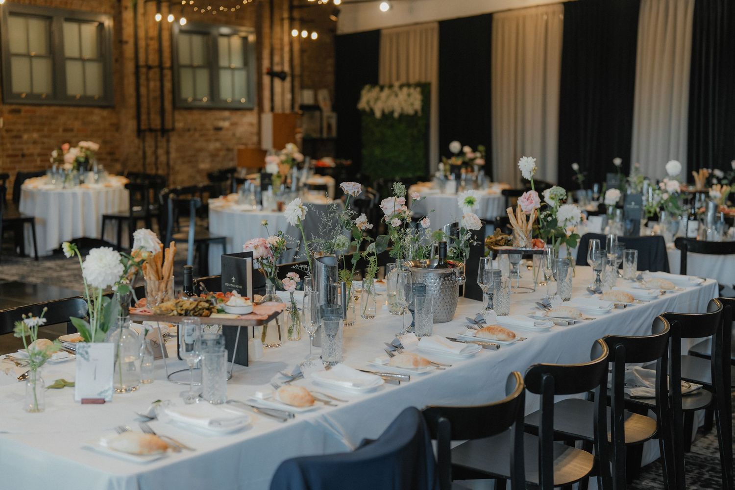 Long banquet tables with elegant place settings and floral centerpieces in a rustic brick-walled reception venue.