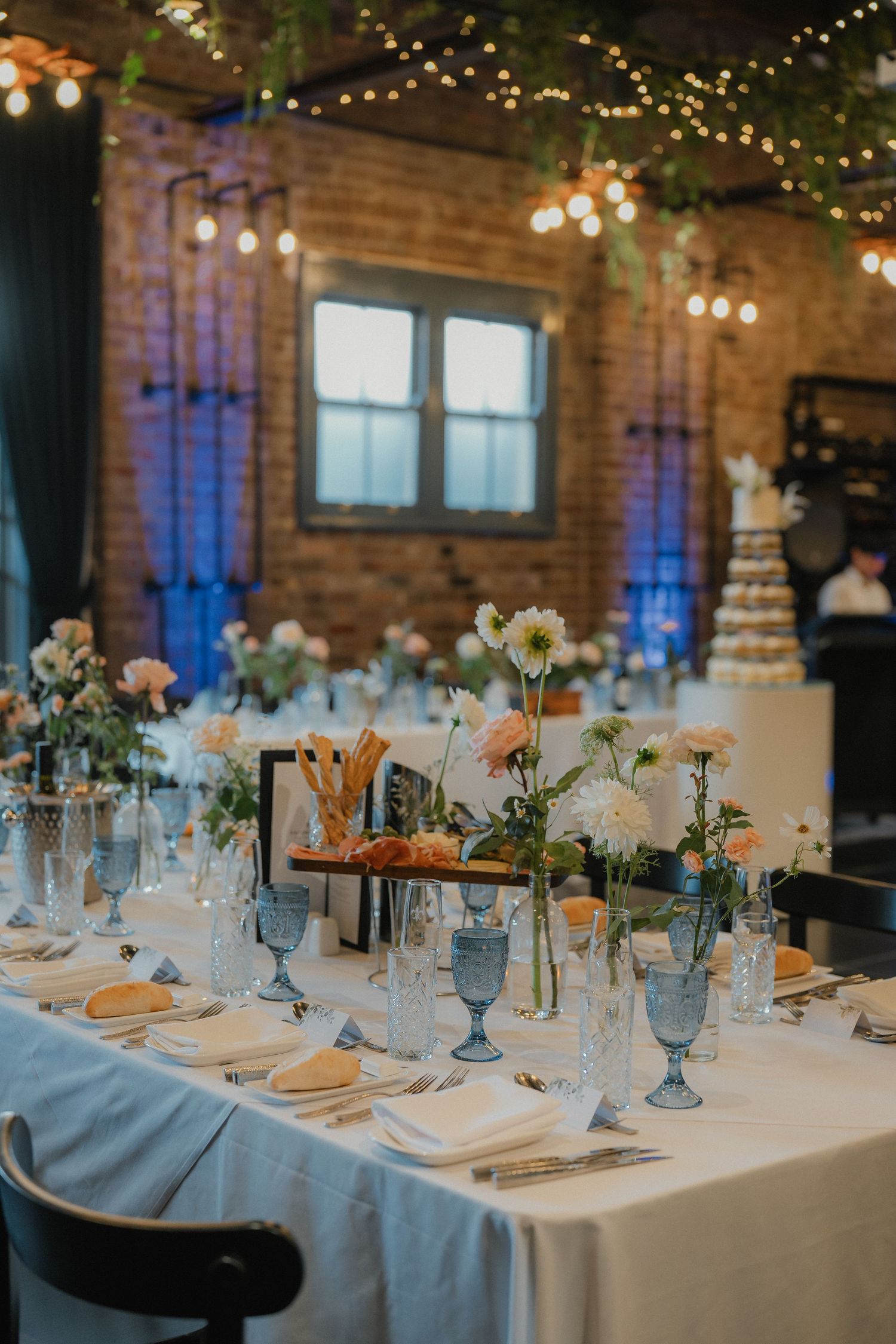 Elegant wedding reception table with blue glassware and floral centerpieces under string lights in rustic venue.