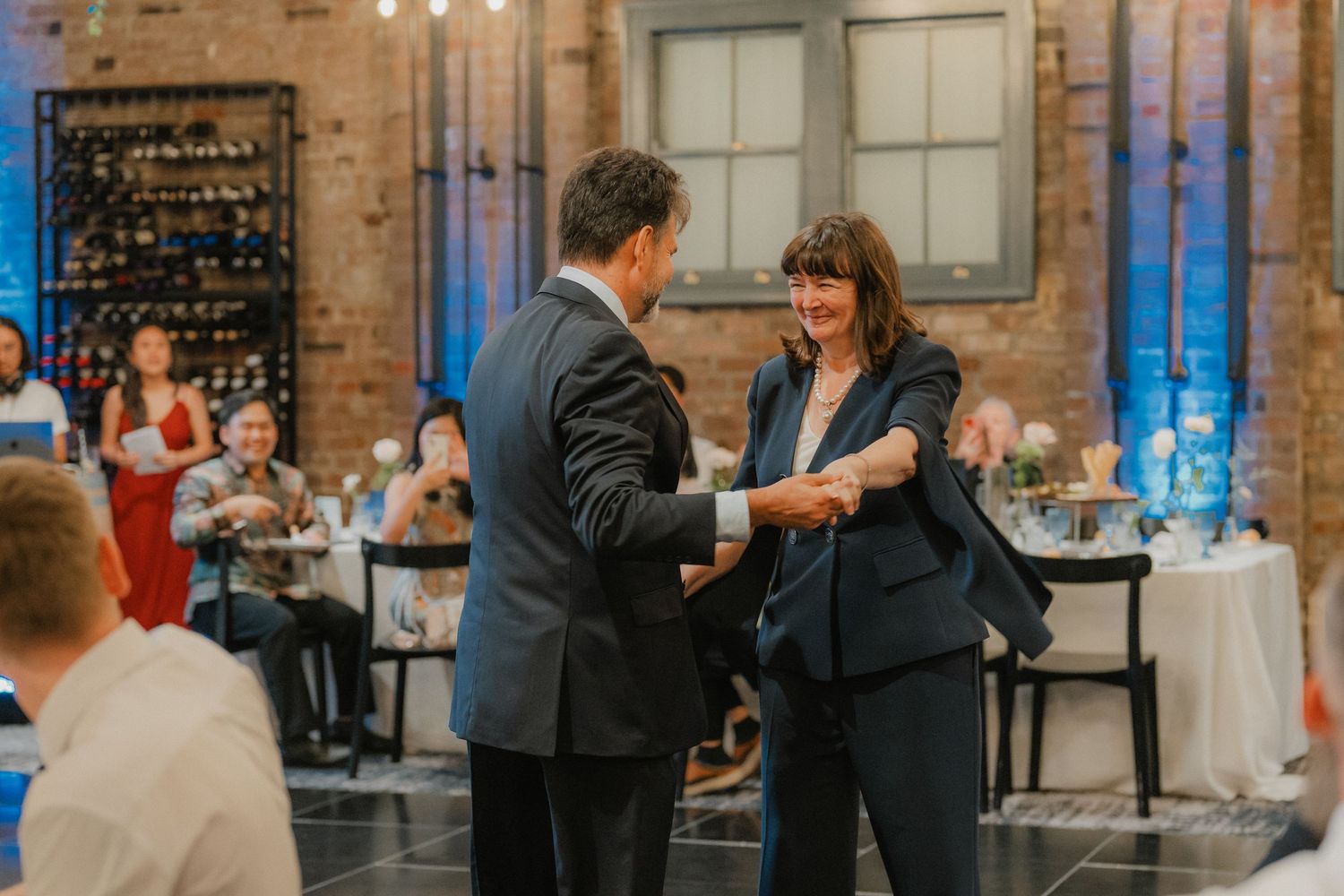 Wedding reception in a rustic brick venue with guests mingling and dancing under warm lighting.